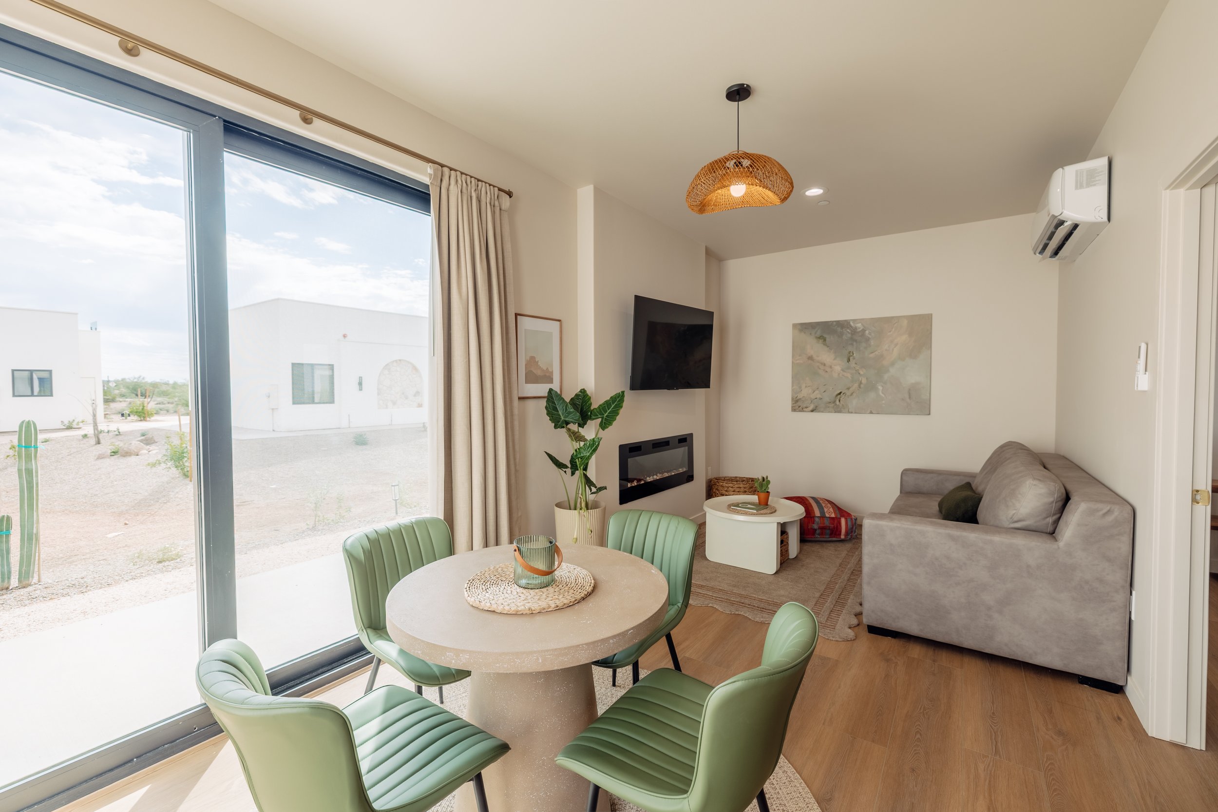 Interior of a modern living room with a dining area, large sliding glass door, green chairs, a round table, a grey sofa, wall art, a wall-mounted TV, and a fireplace.