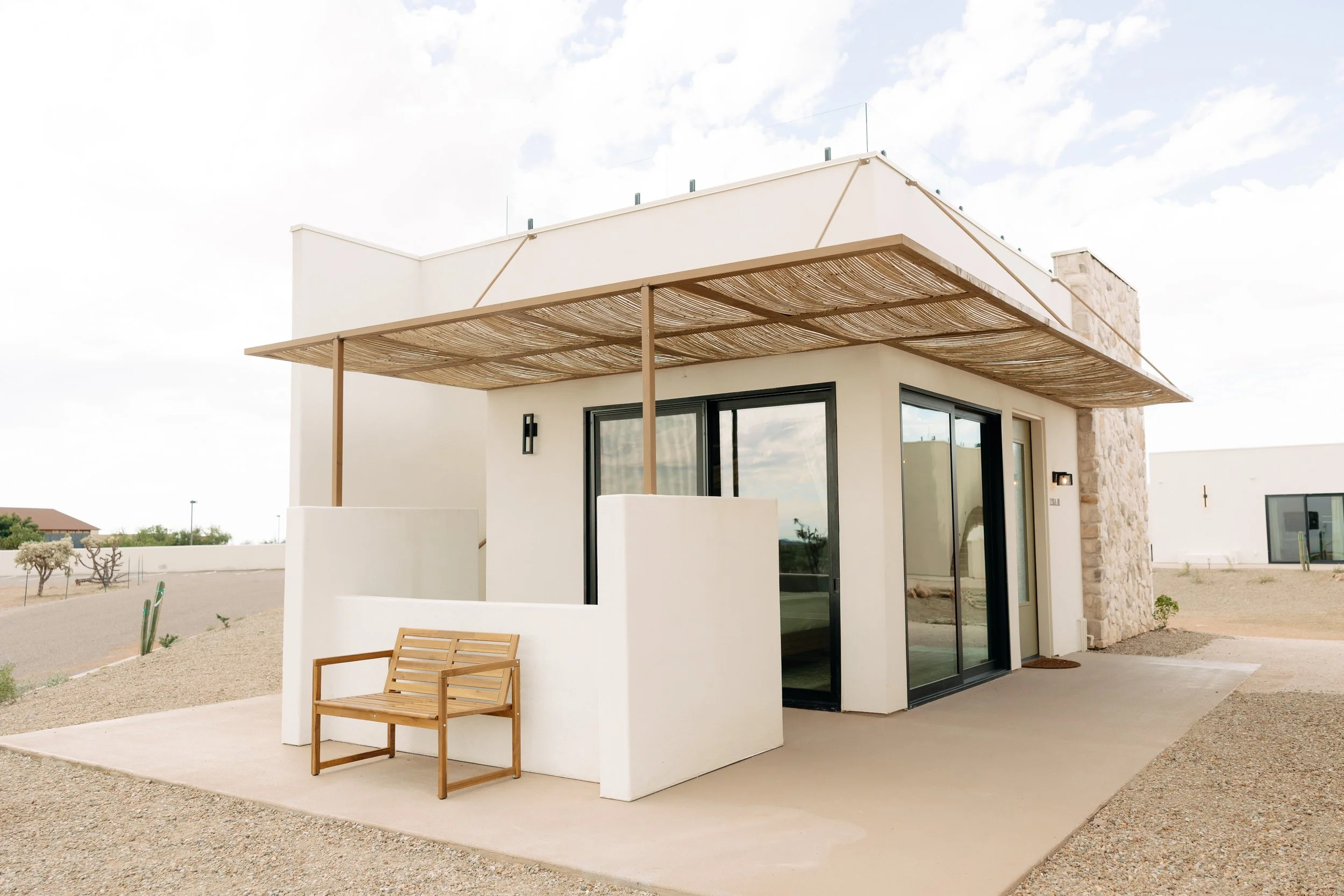 Modern house with white exterior, glass sliding doors, stone wall accents, and a wooden bench on a small patio in a desert landscape.
