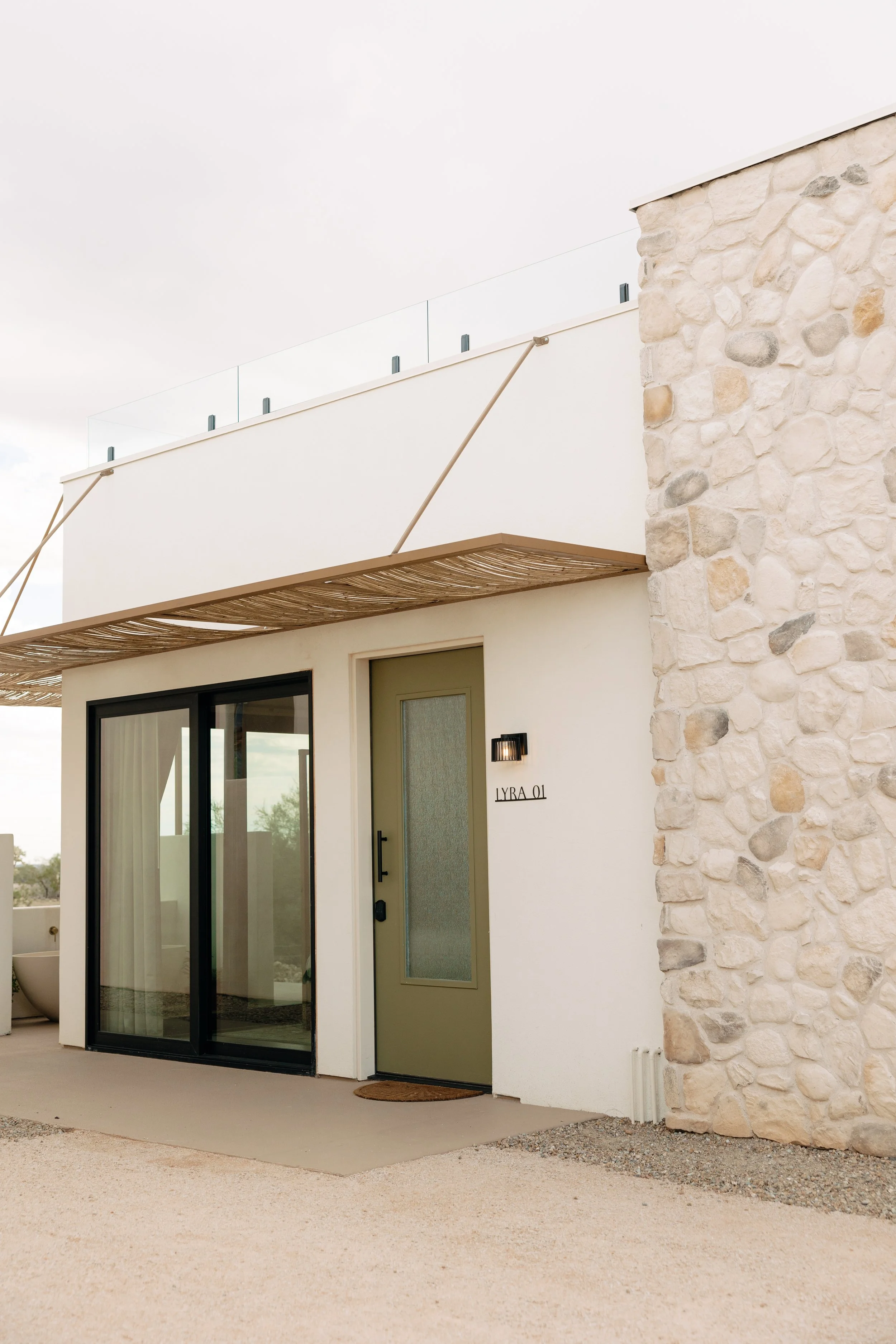 Exterior view of a modern house with a sliding glass door, a green door labeled 'LYRA 01', a stone wall, and a small outdoor light fixture.