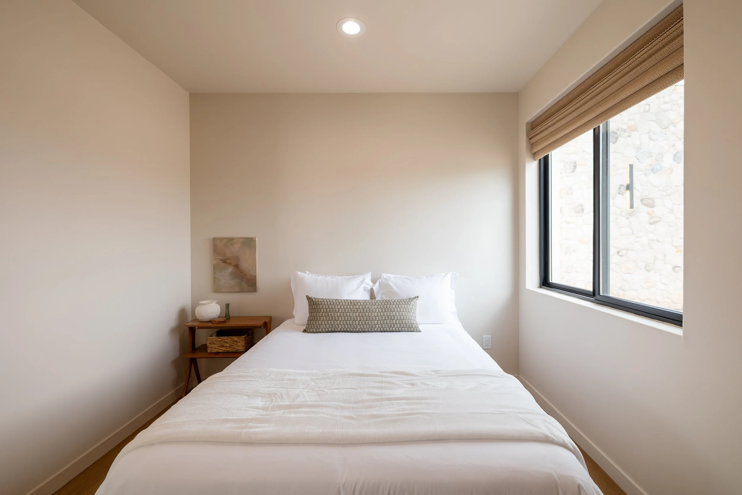 A minimalist bedroom with a white bed, a small wooden side table, a window with a beige shade, and a beige wall art piece.
