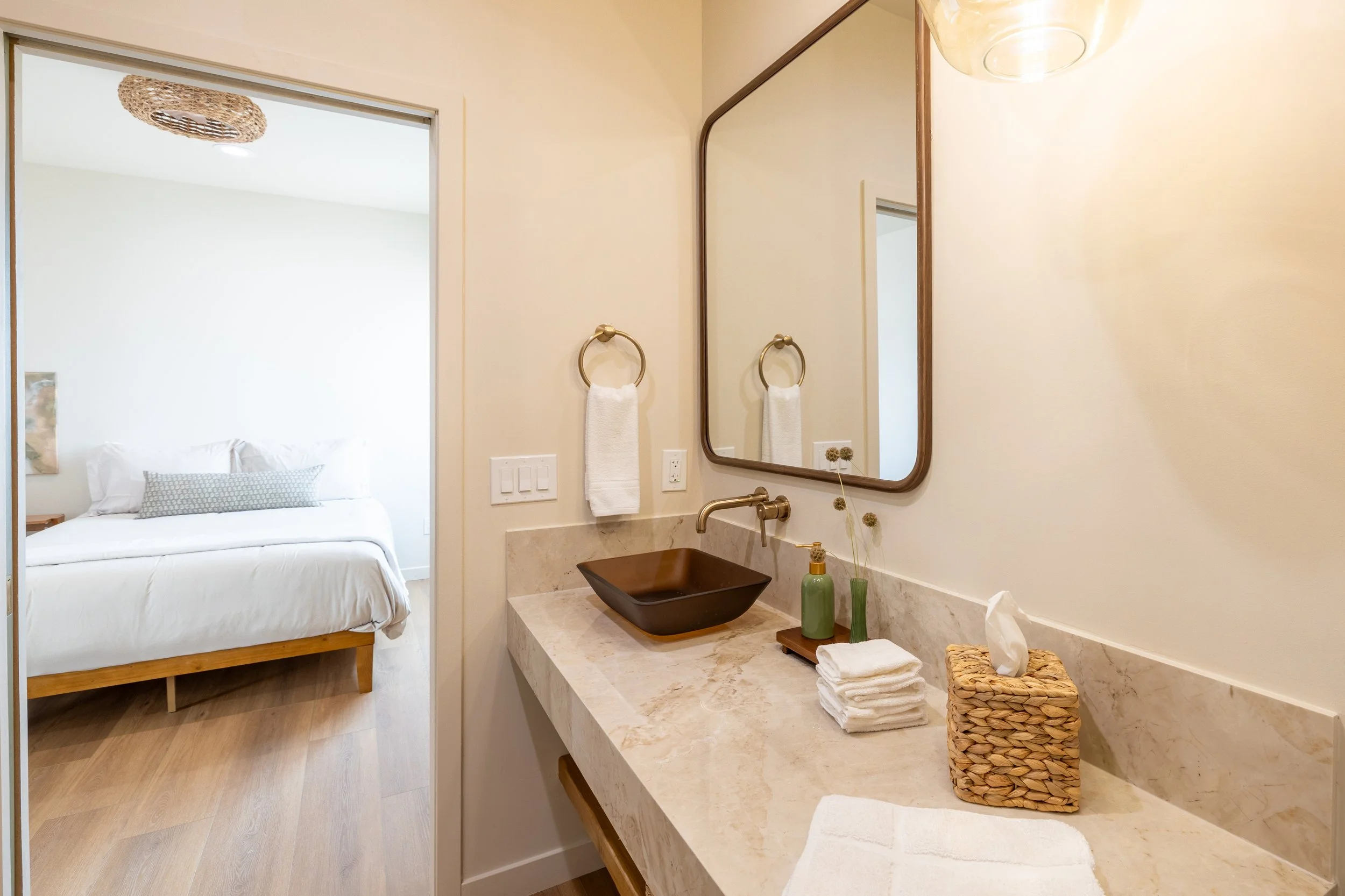 Bathroom vanity with a brown vessel sink, a wall-mounted faucet, a large mirror, a white hand towel on a golden ring, soap, and face towels on the marble counter. Bedroom visible through the doorway with a bed and white linens.