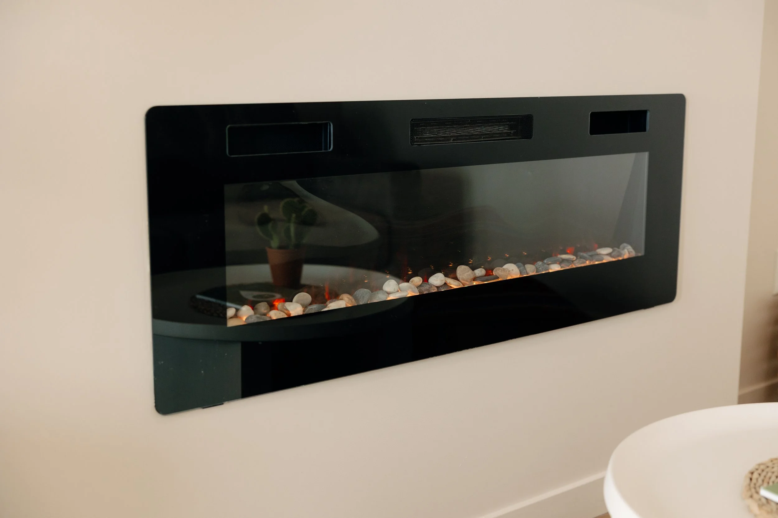 Modern wall-mounted electric fireplace with black trim and stone accents, reflecting a potted plant and furniture nearby.