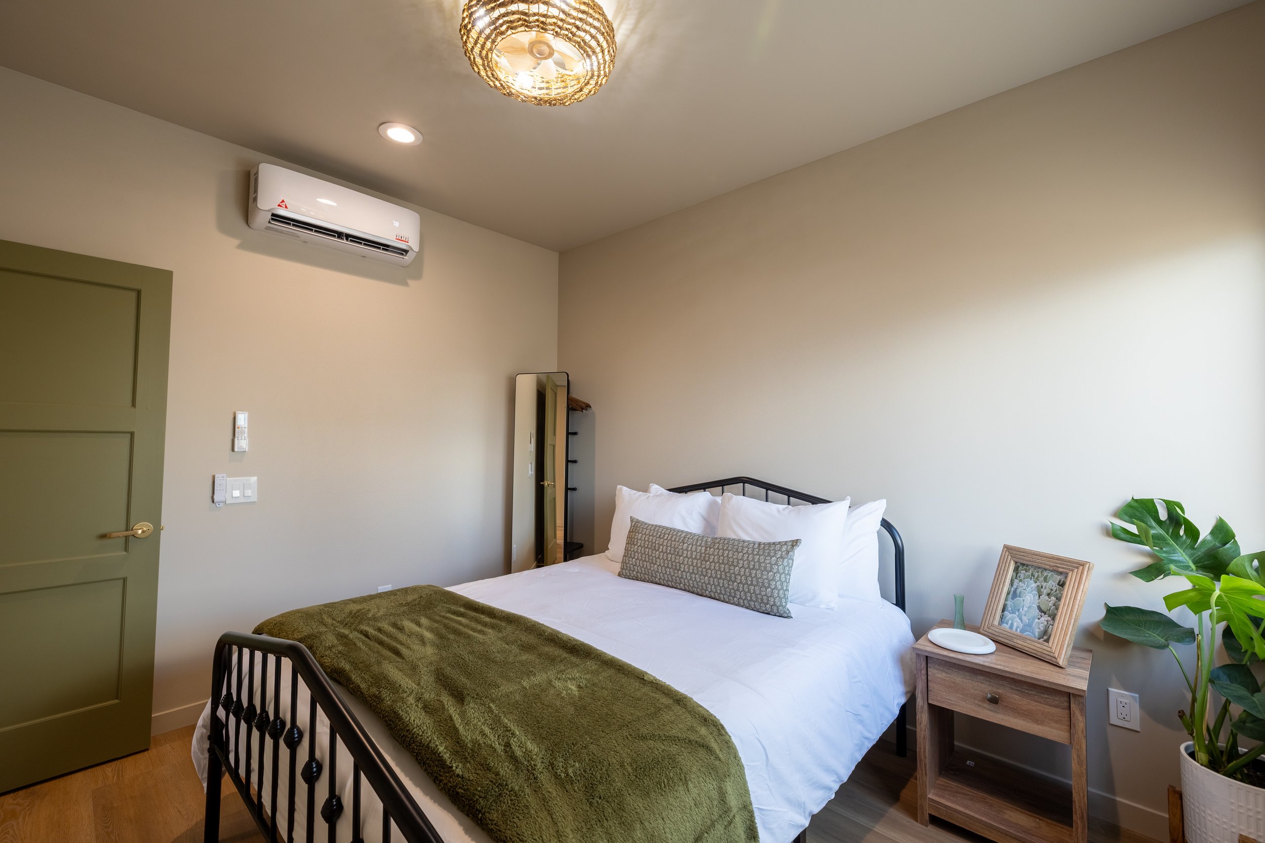 Bedroom with a bed, pillows, a nightstand with a framed photo, a plant, and a mirror, light beige walls, and a ceiling light.