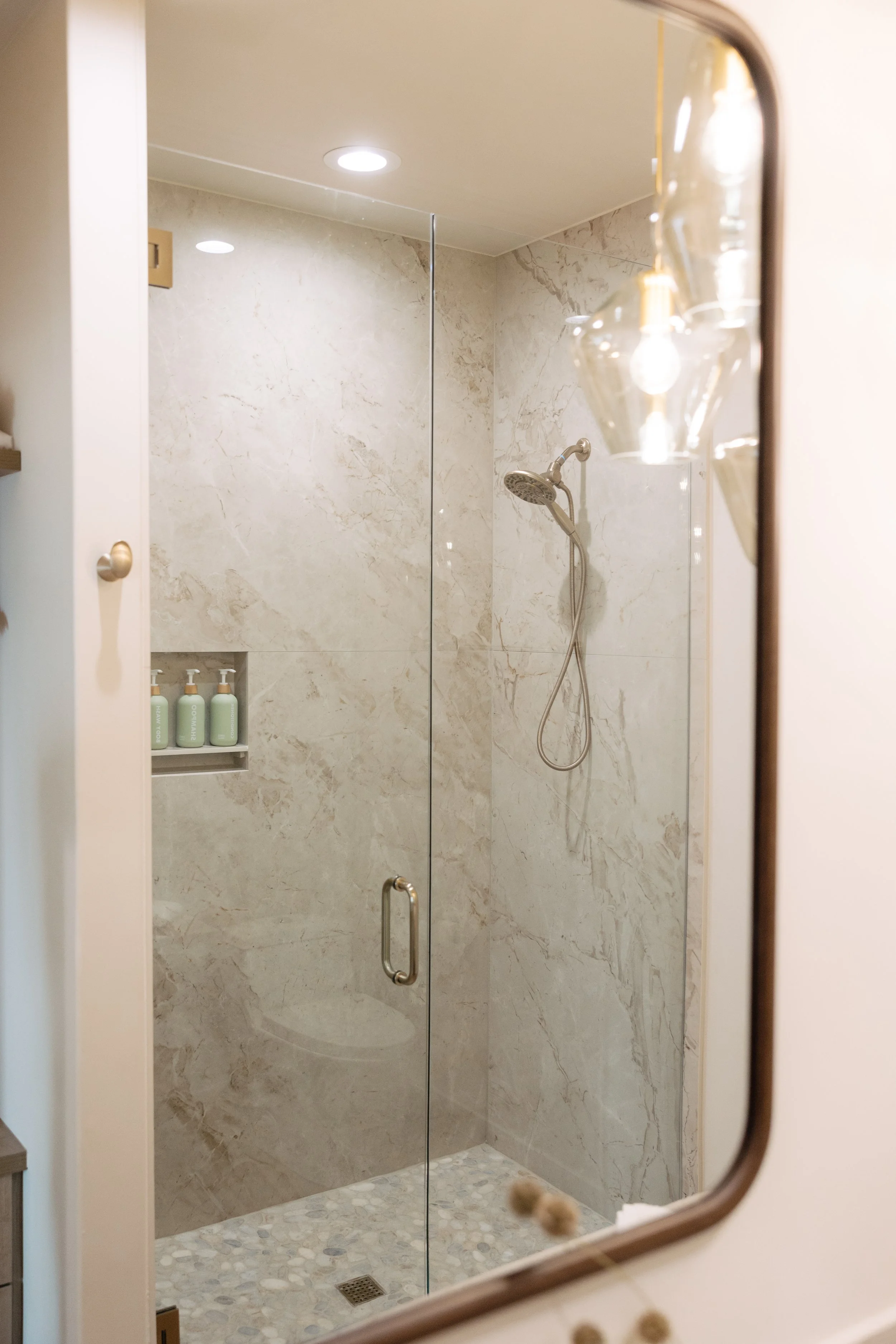 A glass-enclosed shower with a marble interior, featuring a handheld showerhead, a recessed shelf with toiletries, and pebble tile flooring, reflected in a mirror.