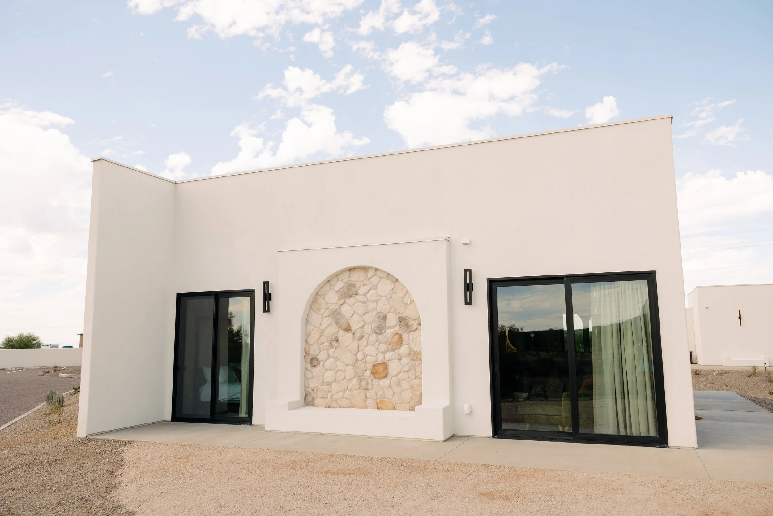 A modern, white, rectangular house with large sliding glass doors, an arched stone accent wall, and simple black exterior wall lights, set against a blue sky with scattered clouds.