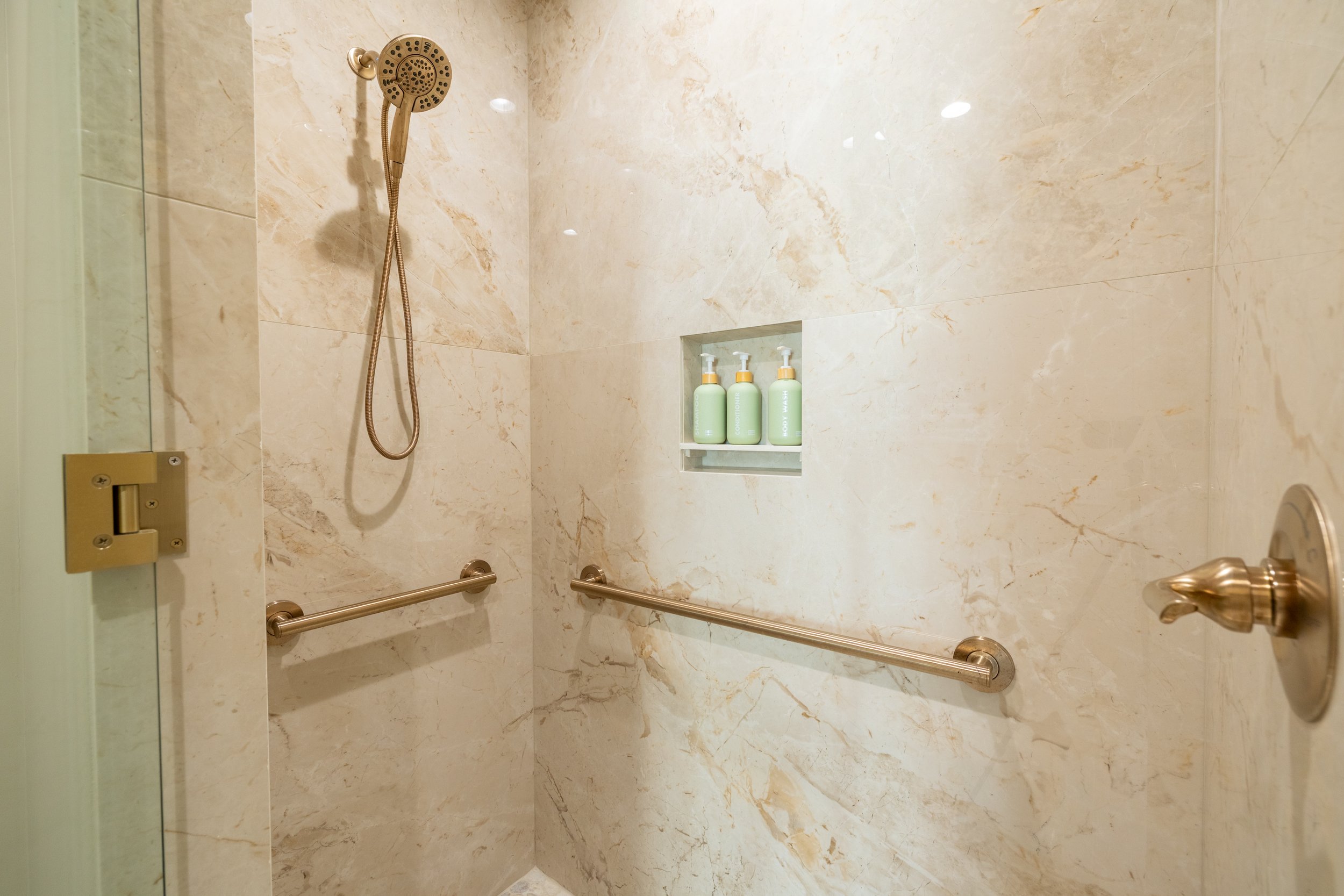 Shower with beige marble tiles, a wall-mounted showerhead, and two horizontal grab bars. There is a built-in niche with three bottles of shampoo or body wash.