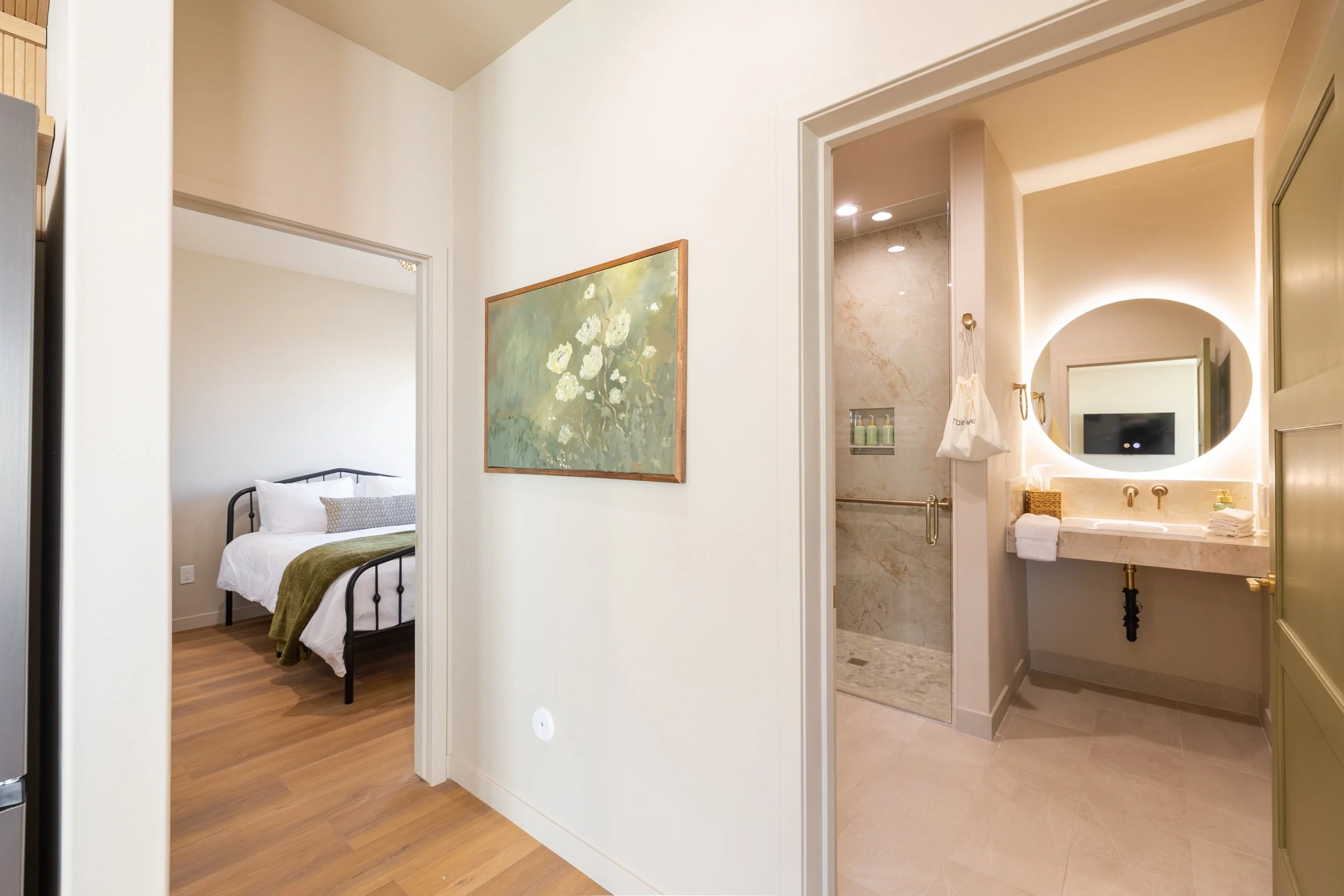 View of a hotel or apartment interior with a bedroom to the left and a bathroom to the right, including a shower and sink with a mirror.