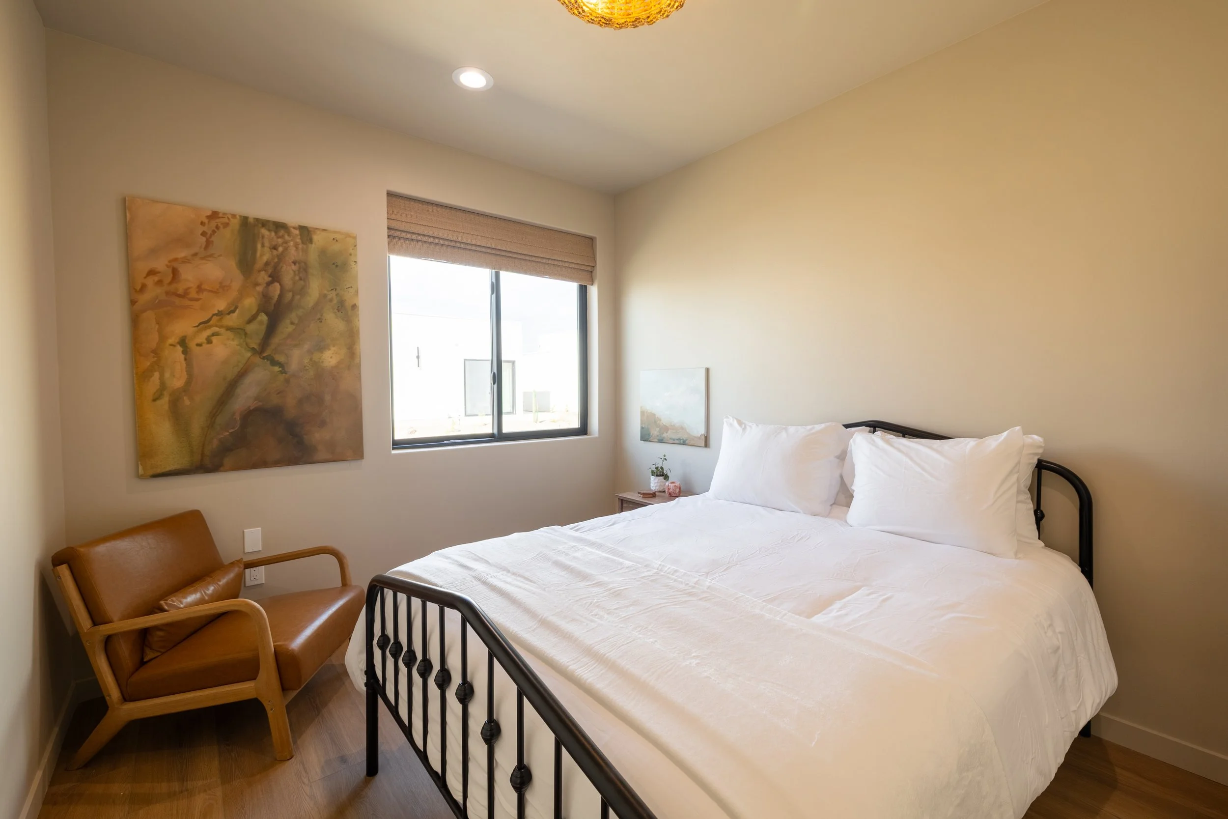 A bedroom with a black metal bed frame covered in white bedding and pillows, a small wooden nightstand with a potted plant and decorative item, a brown armchair with a cushion, and artwork on the walls. There is a window with a beige shade letting in