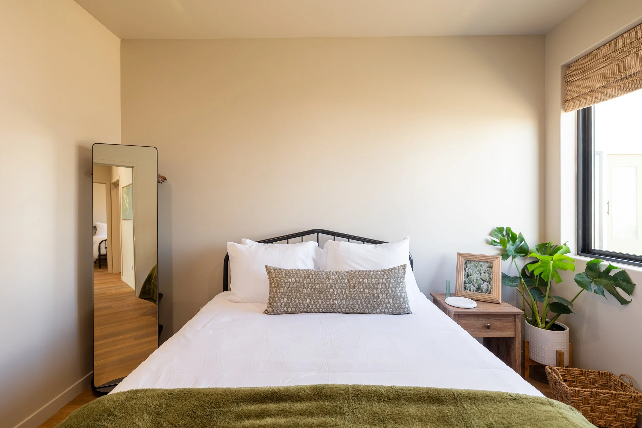 A bedroom with a bed, a mirror, a nightstand, a framed picture, a large plant, a window, and a wicker basket.