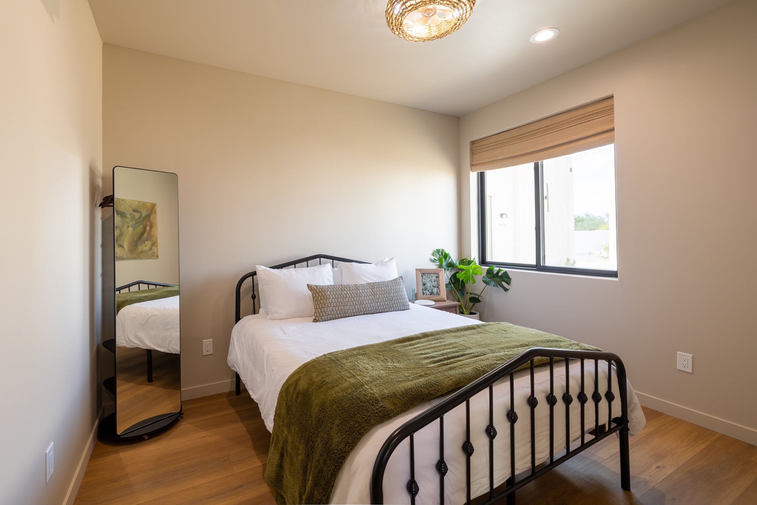 Bedroom with a black metal bed, white bedding, green throw blanket, window with a beige shade, nightstand with a plant, framed photo, and a mirror in the corner.