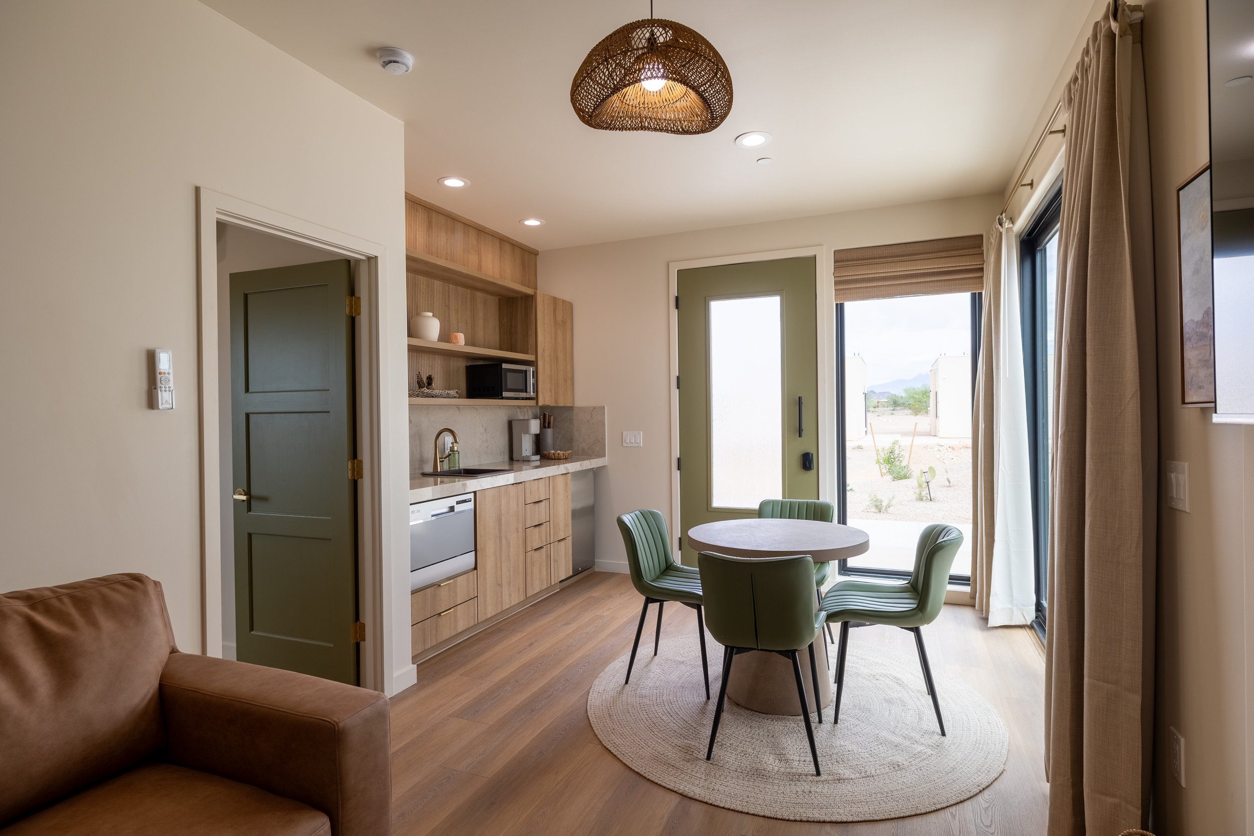 Interior view of a modern, sunlit kitchen and dining area with a round table and six green chairs, a kitchenette with wood cabinets, and sliding glass doors leading outside.