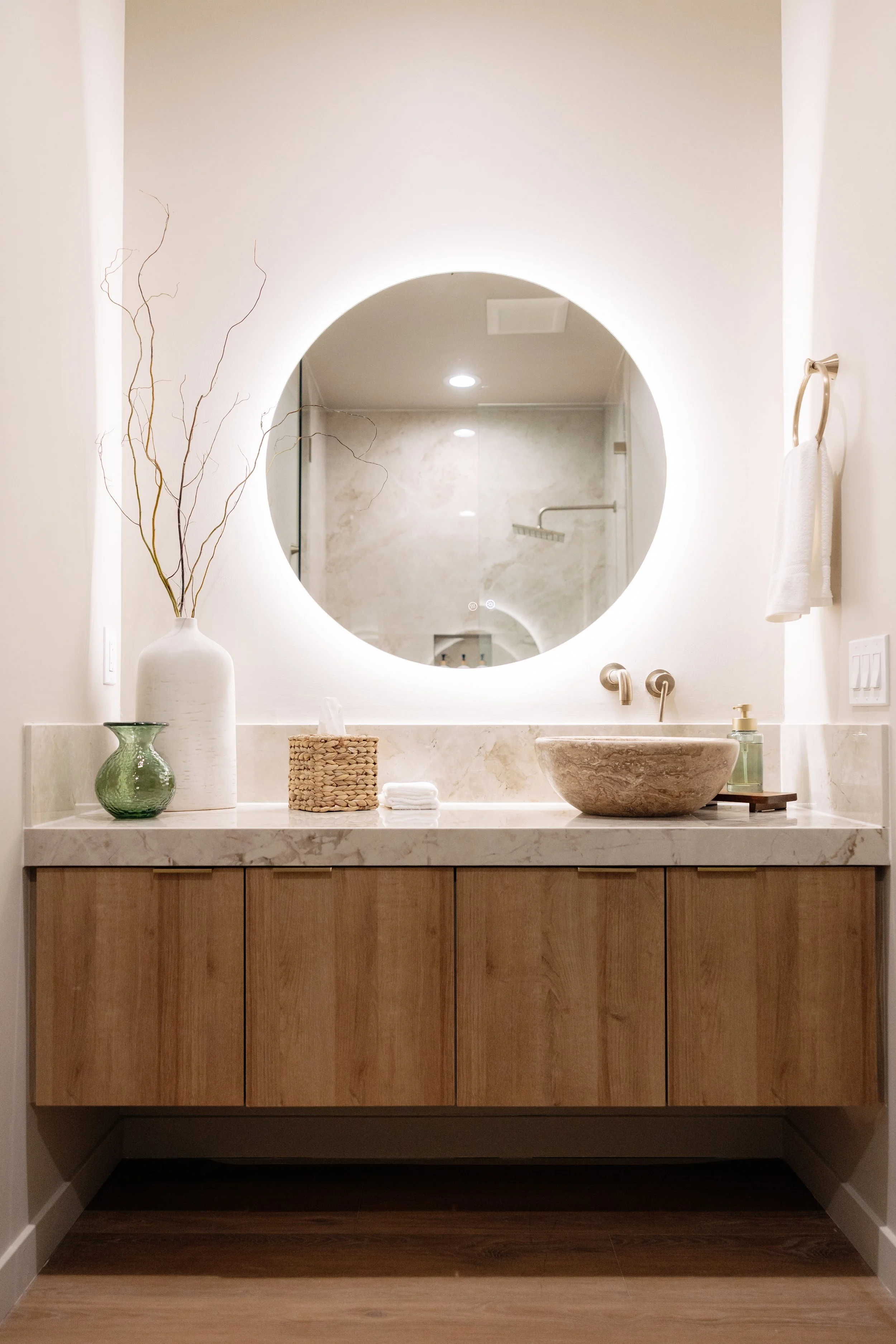 Bathroom vanity with a round backlit mirror, a stone vessel sink, a wooden cabinet, a white vase with dried branches, a green decorative glass, a woven tissue box holder, and a small soap dispenser.