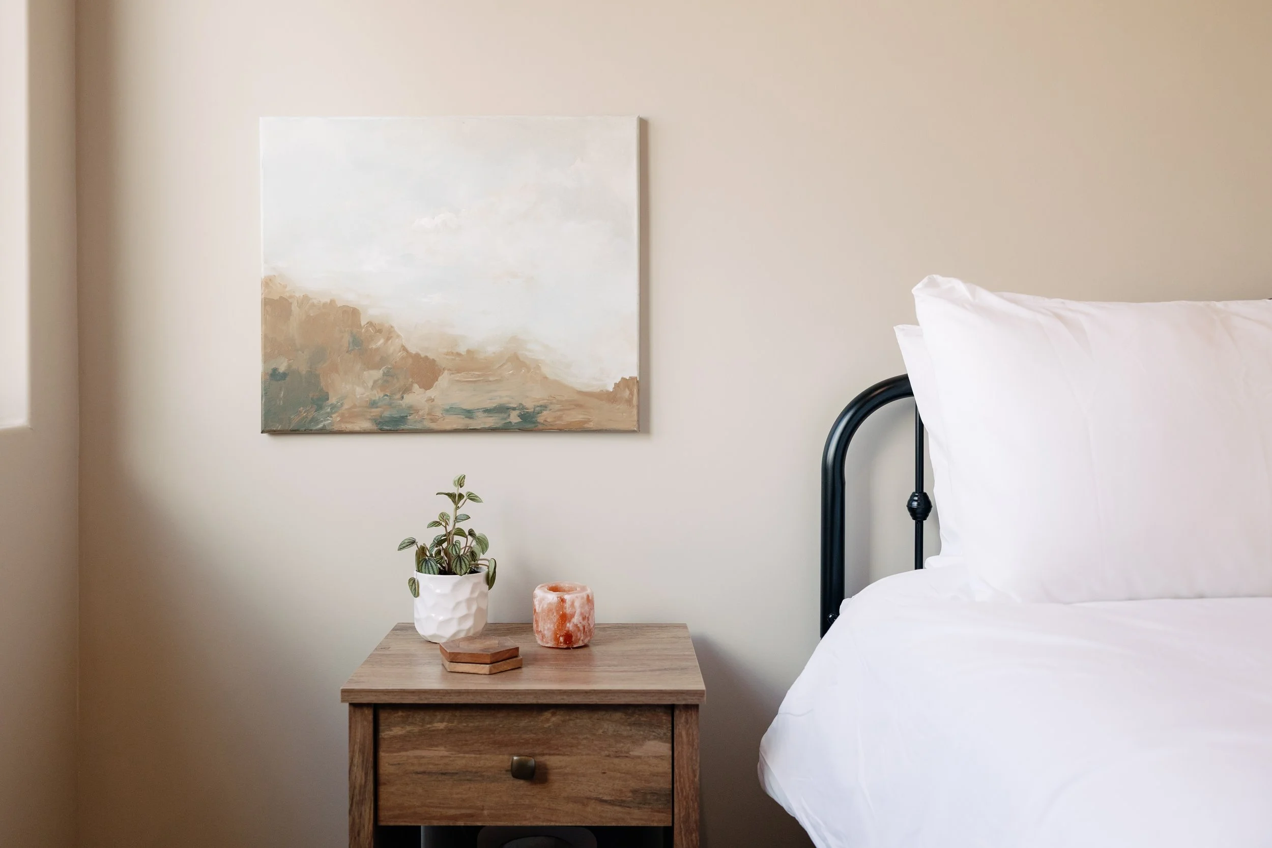 A bedroom corner with a wooden nightstand, a white potted plant, salt lamp, and a canvas painting on a cream wall. Part of a bed with white bedding and a black headboard is visible.