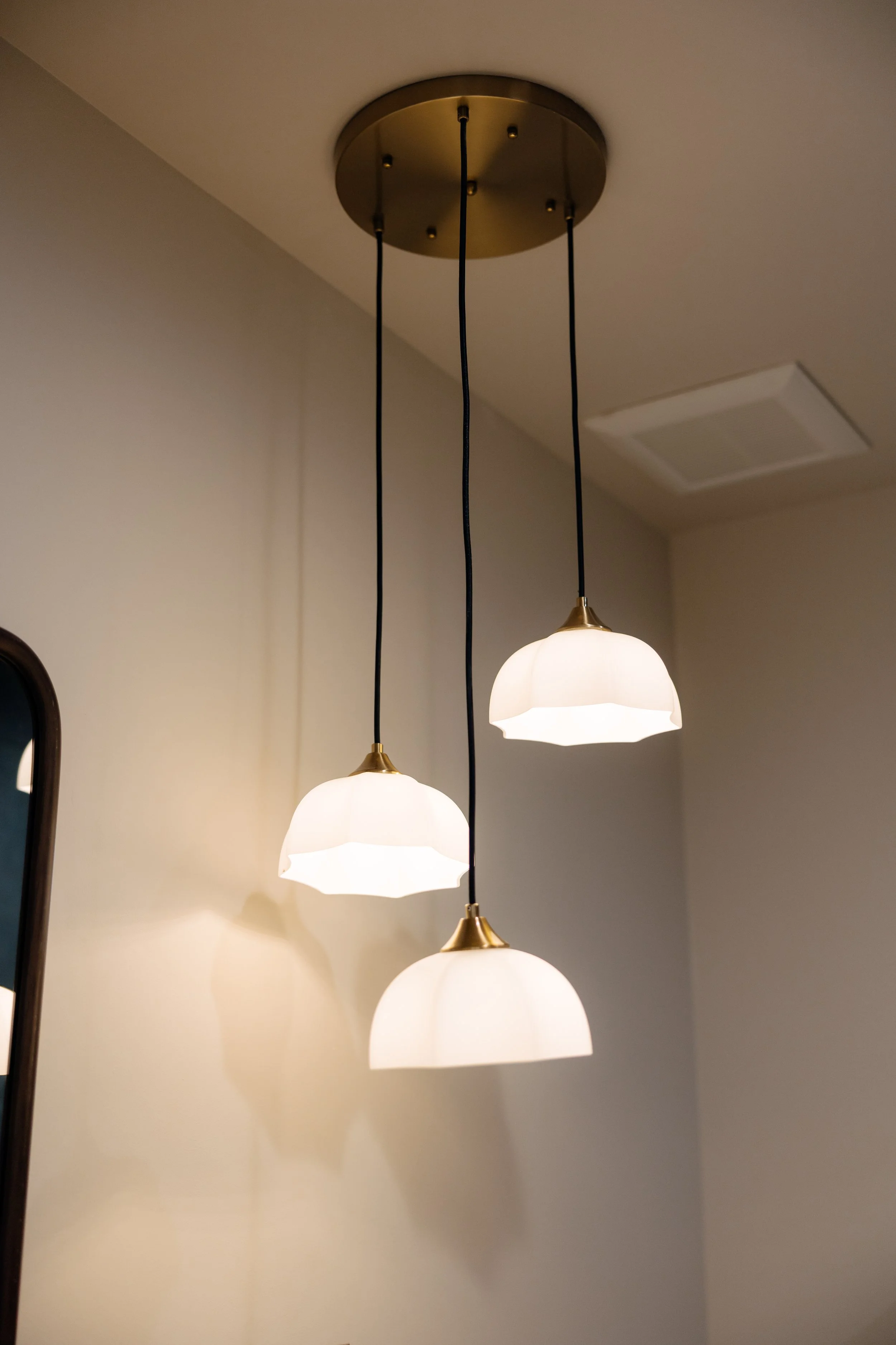 A modern pendant light fixture with three white, semi-flush glass shades hanging from black cords, attached to a round brass ceiling mount.
