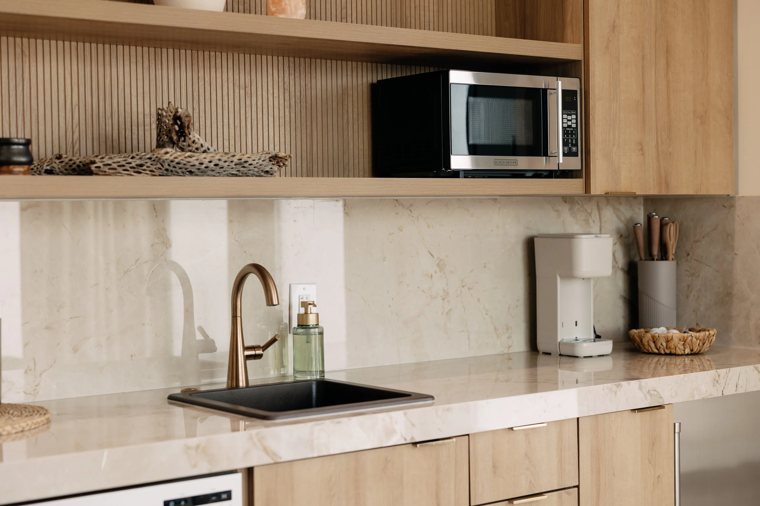 Kitchen with wooden cabinets, marble countertop, black sink, gold faucet, soap dispenser, microwave, coffee maker, utensil holder, and decorative items.