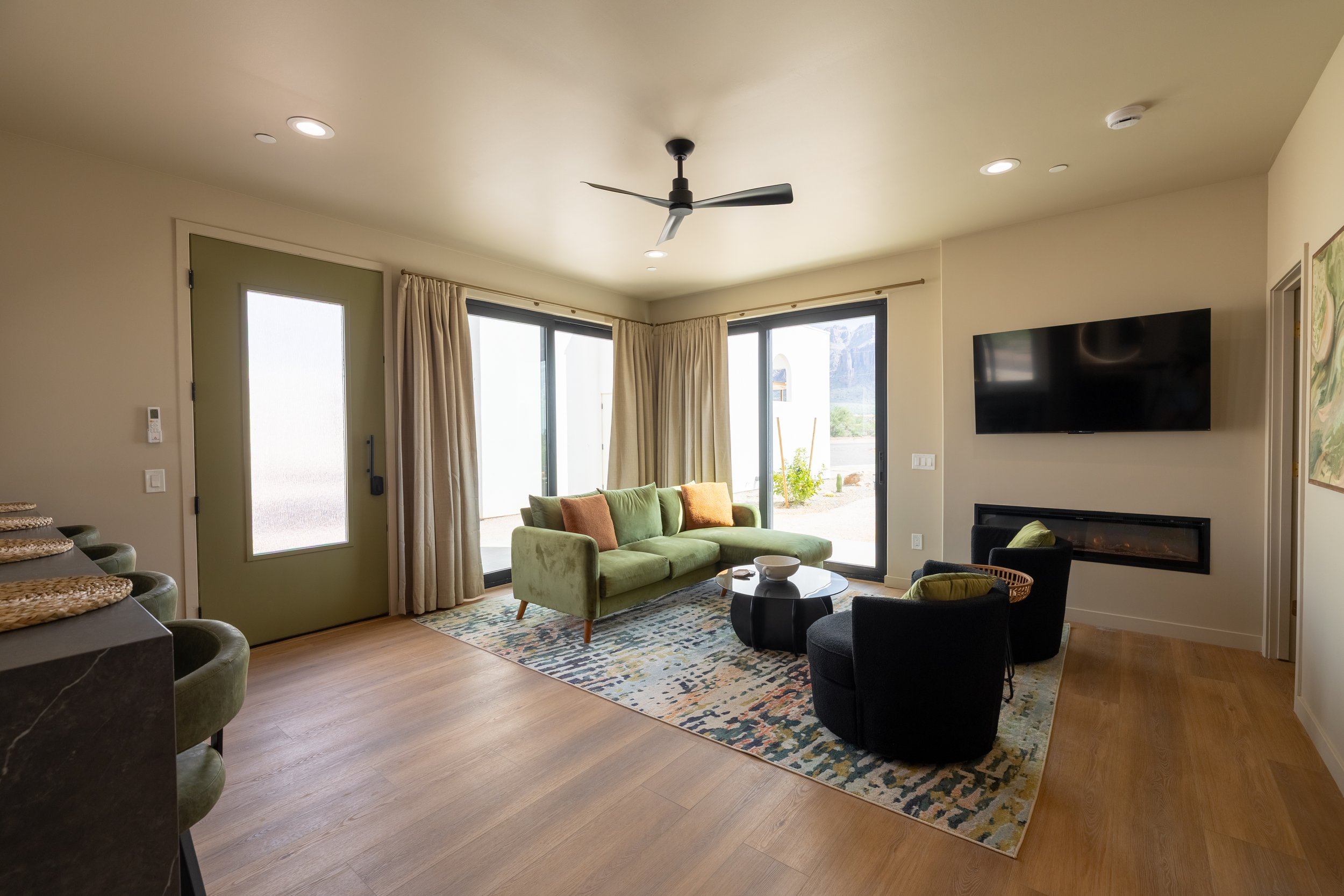 Living room with green sofa, black chairs, TV on wall, sliding glass doors with curtains, and a patterned rug.