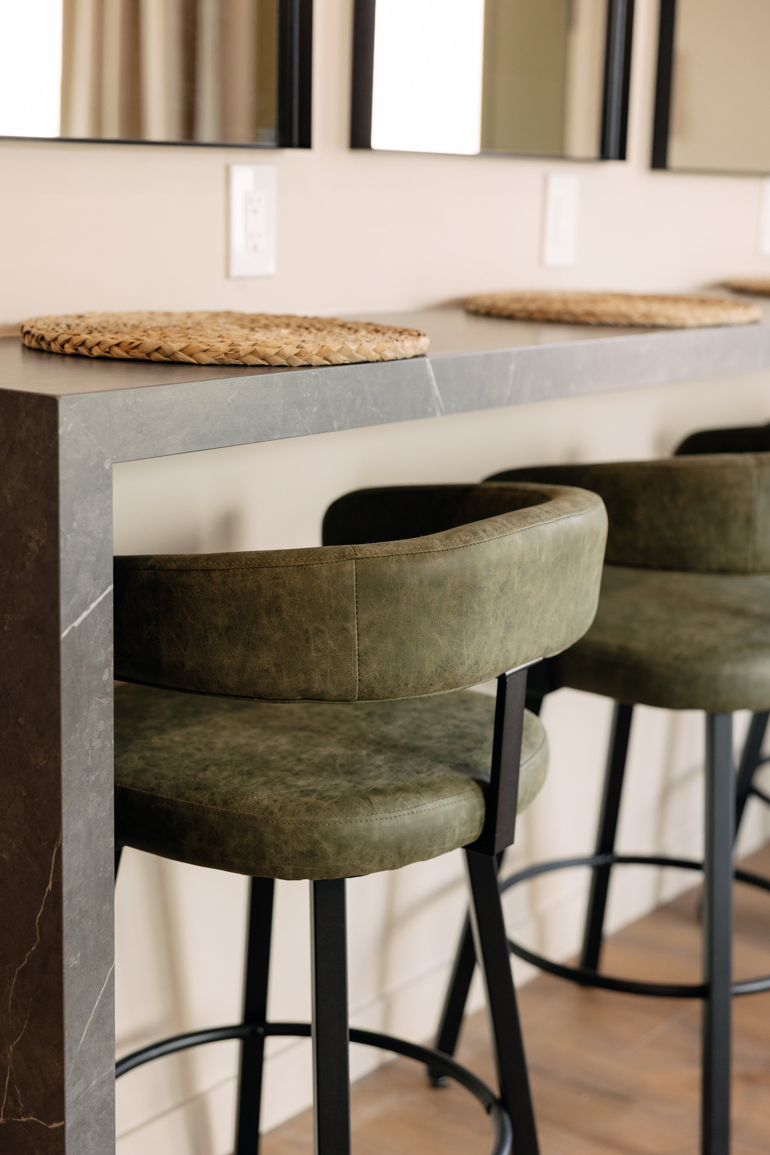 Close-up of a kitchen or bar area with a gray stone counter, green upholstered bar stools with black metal legs, and woven placemats on the counter; mirrors and electrical outlets on the wall in the background.