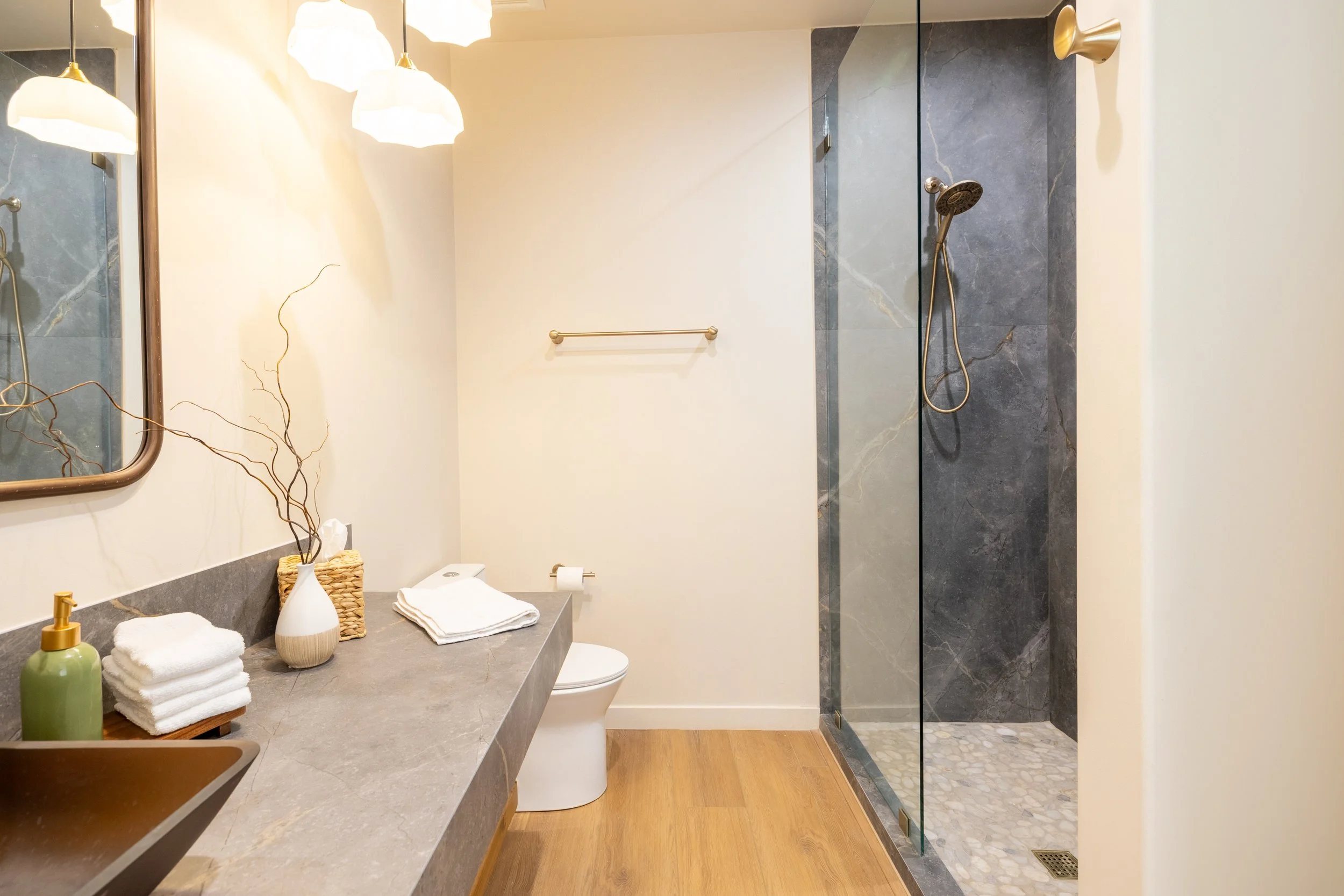 Modern bathroom with a walk-in shower featuring dark gray stone tiles and pebble flooring, a white toilet, and a gray countertop with neatly folded towels, a soap dispenser, a decorative vase with branches, and a mirror. Warm lighting fixtures hang f