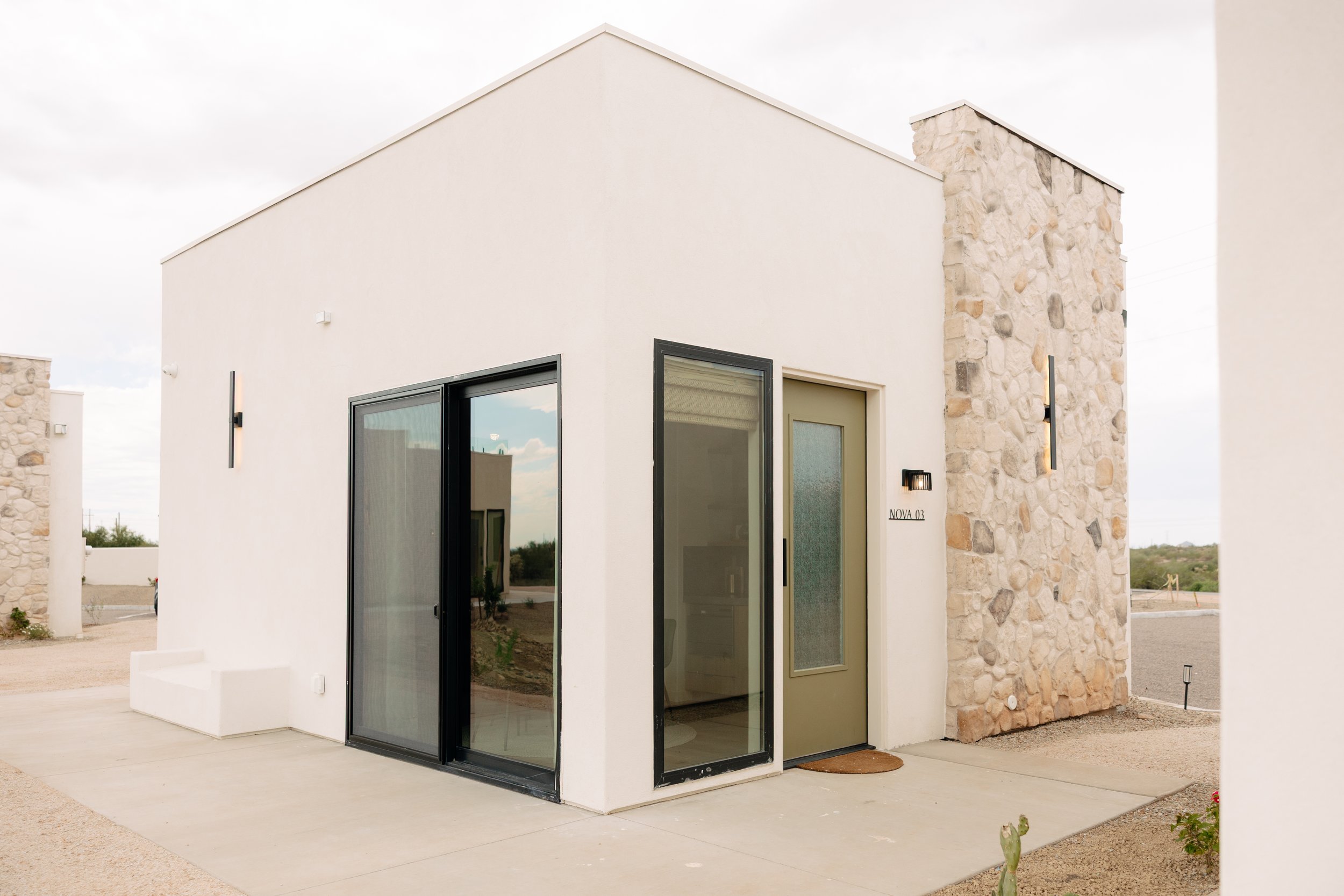 Modern house with white walls, stone accents, and a wooden overhang, surrounded by desert landscaping including cacti, rocks, and drought-tolerant plants, under a blue sky with scattered clouds.