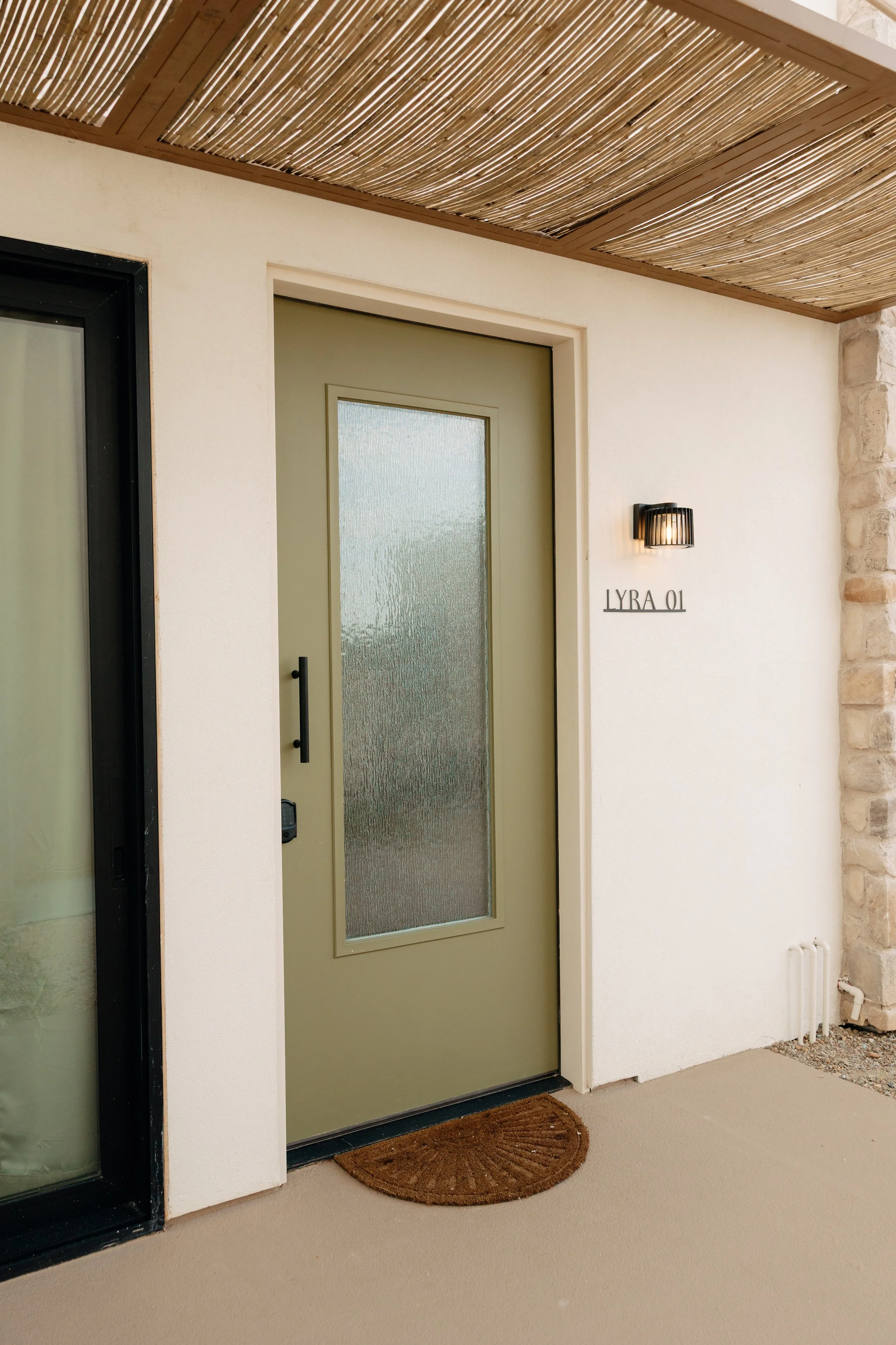 Front door with a frosted glass panel, a black handle, and a brown doormat in front. To the right, a wall-mounted black light fixture and a sign that reads 'LYRA 01'. An exterior wall with a brick column and a bamboo ceiling above.