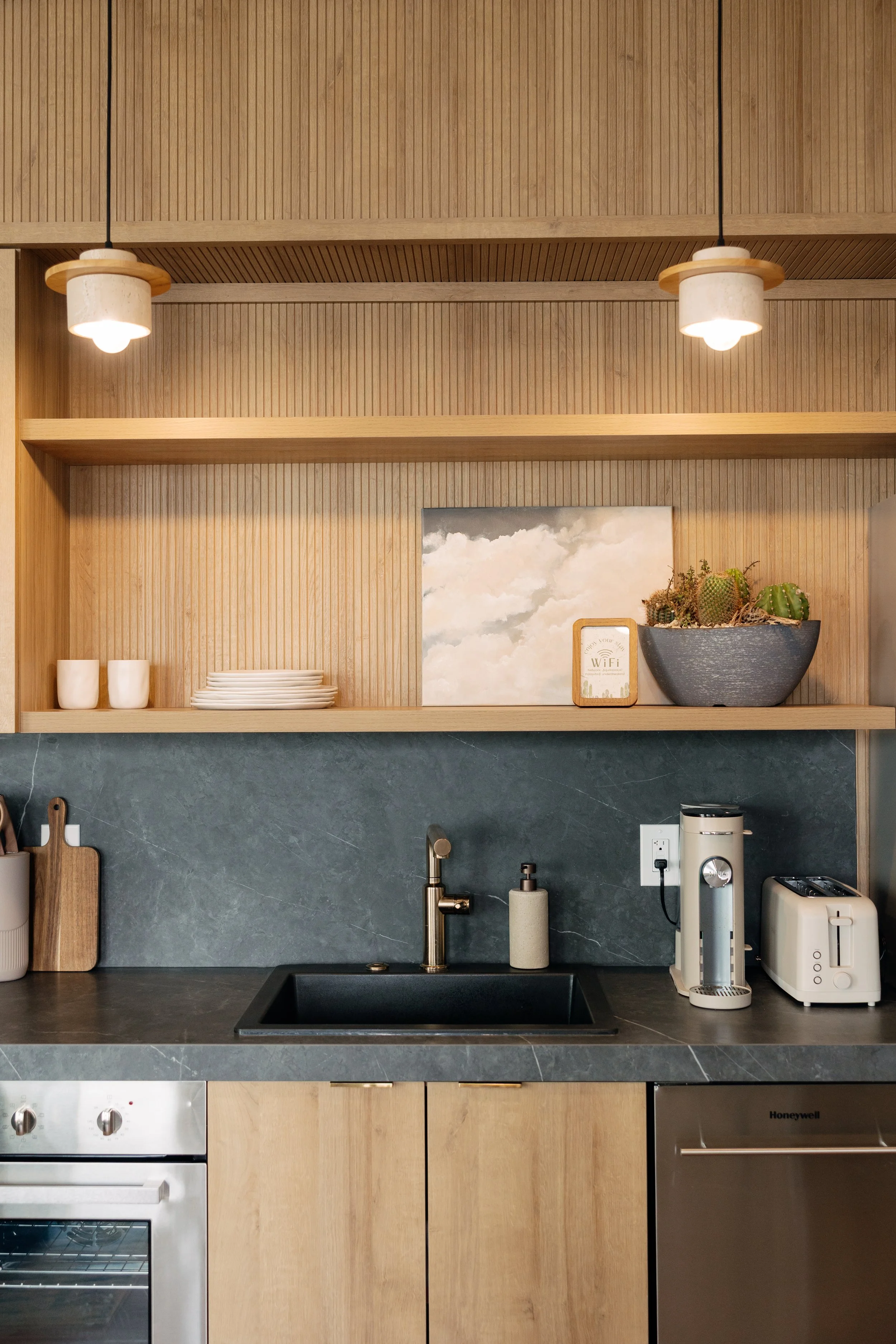 Modern kitchen with wooden cabinets, a grey marble countertop, and a black sink. Open shelves hold some white dishes, cups, and a potted cactus. Small appliances like a toaster and a coffee machine are on the counter.
