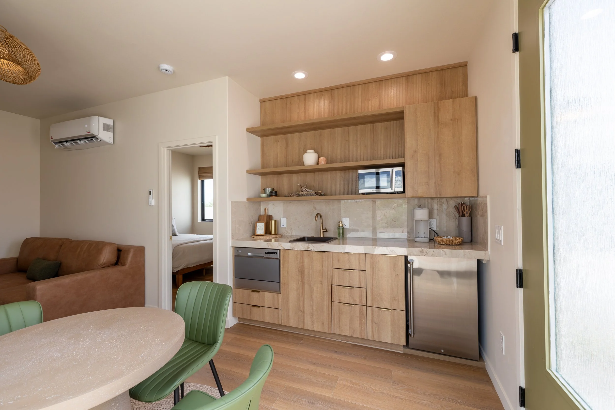 Modern kitchen with wooden cabinets, marble countertops, and open shelving in a living space with a dining table and chairs, adjacent to a bedroom visible through an open door.