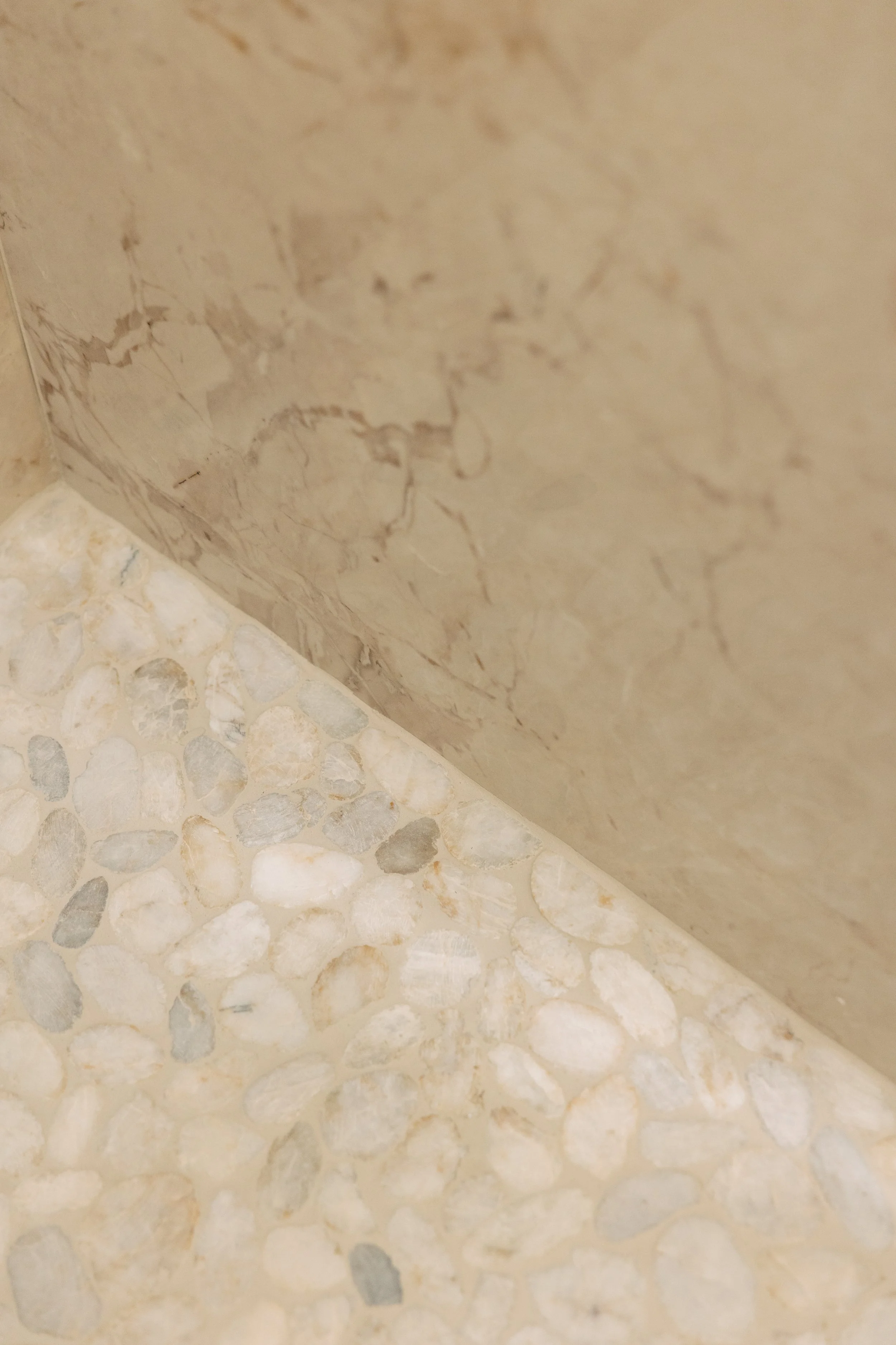 Close-up photo of a marble wall with a beige and brown pattern and a tiled floor with small rounded white and gray stones.