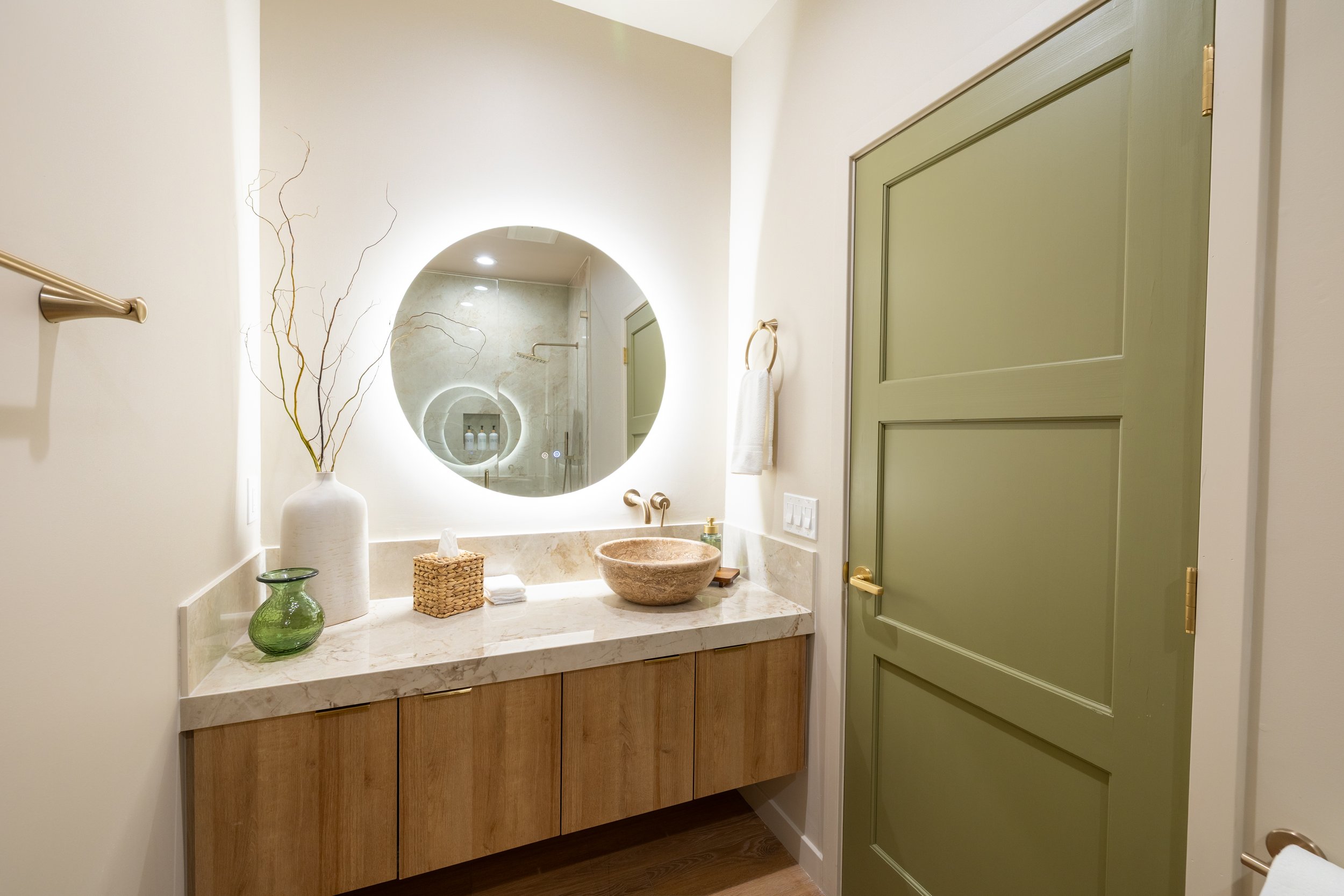 Modern bathroom with a marble countertop, stone vessel sink, circular mirror, and green door.