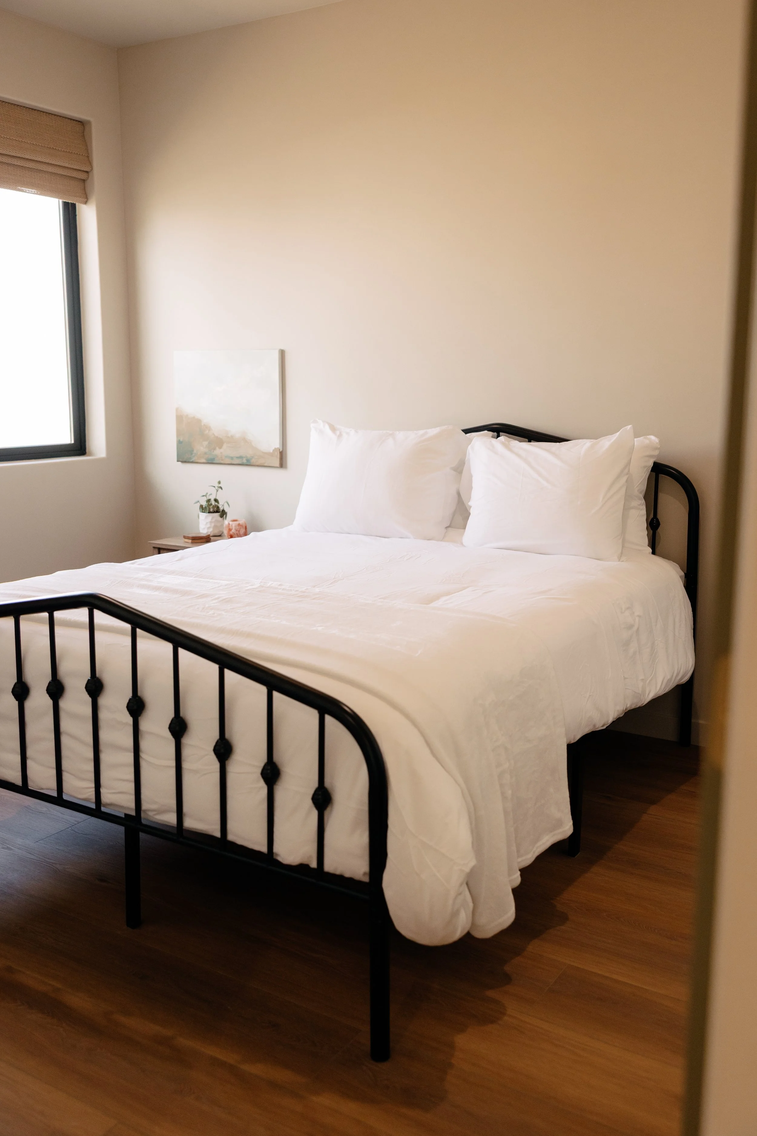 A neatly made bed with white bedding and pillows in a bedroom with a wooden floor, a side table with a small plant and a painting, and a window with a blind.