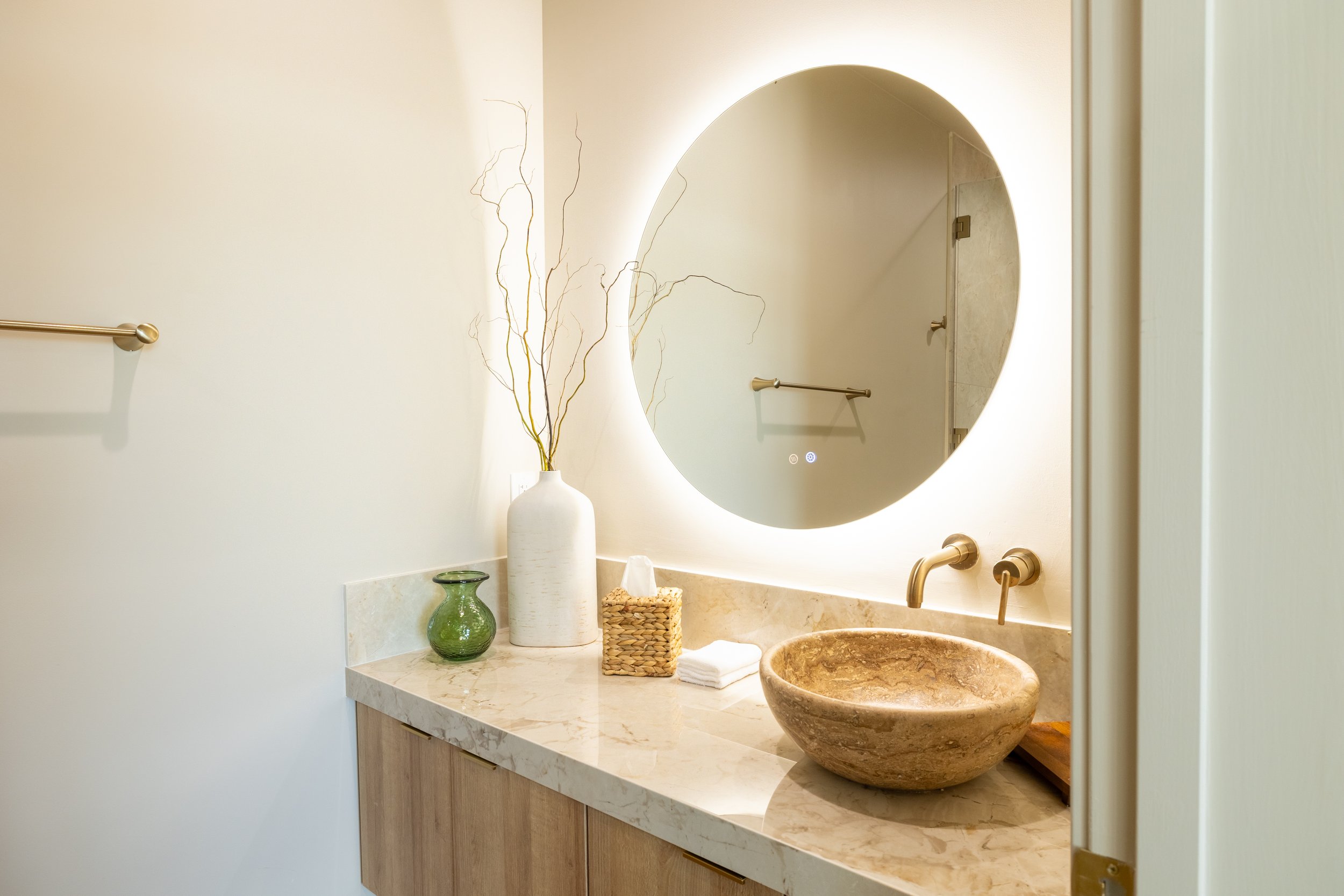 Bathroom vanity with round mirror, stone vessel sink, gold faucet, decorative vases, tissue box, and towel.