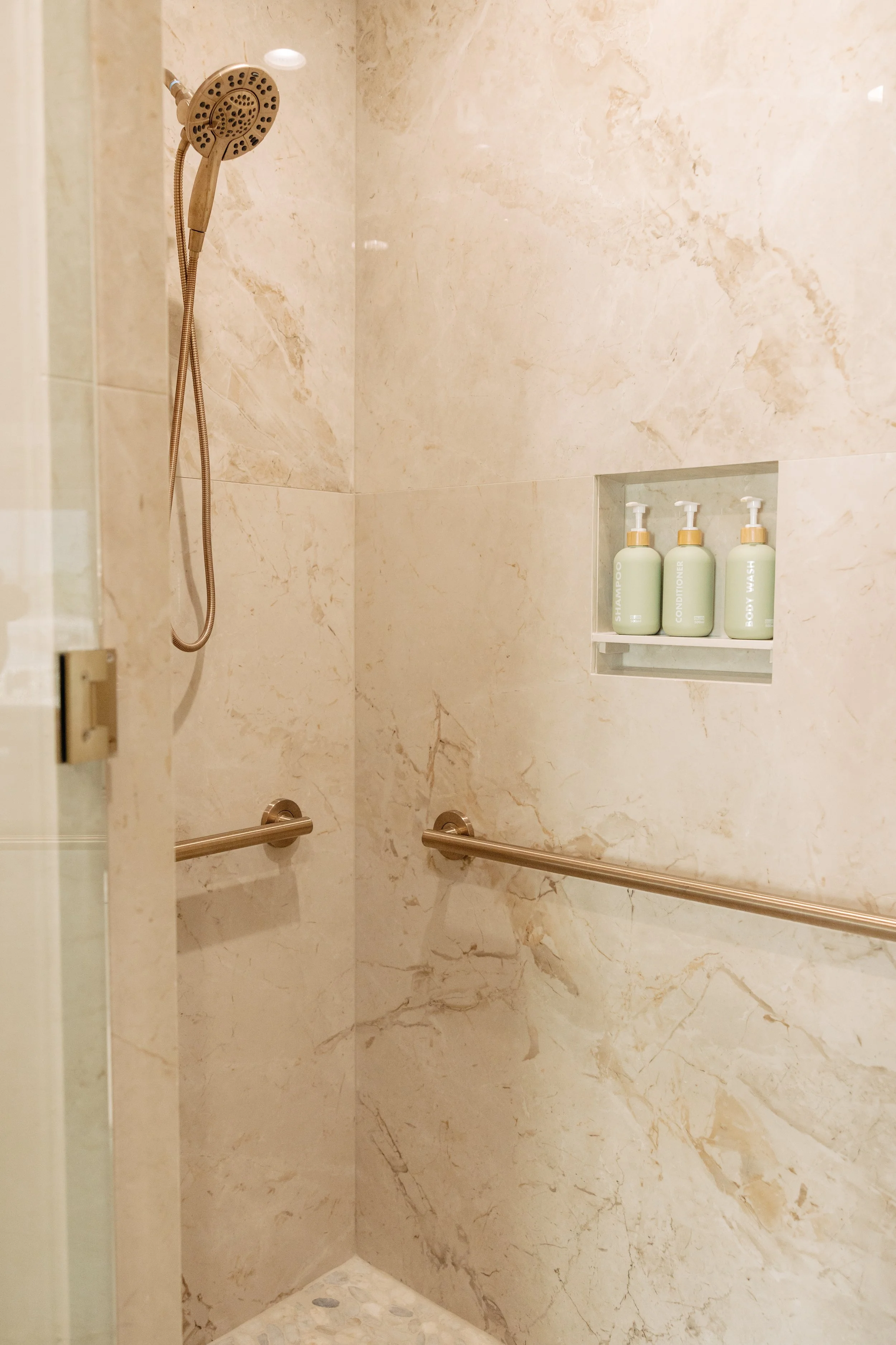 Shower area with beige marble walls, handheld showerhead, safety grab bars, and built-in shelf with shampoo, conditioner, and body wash bottles.