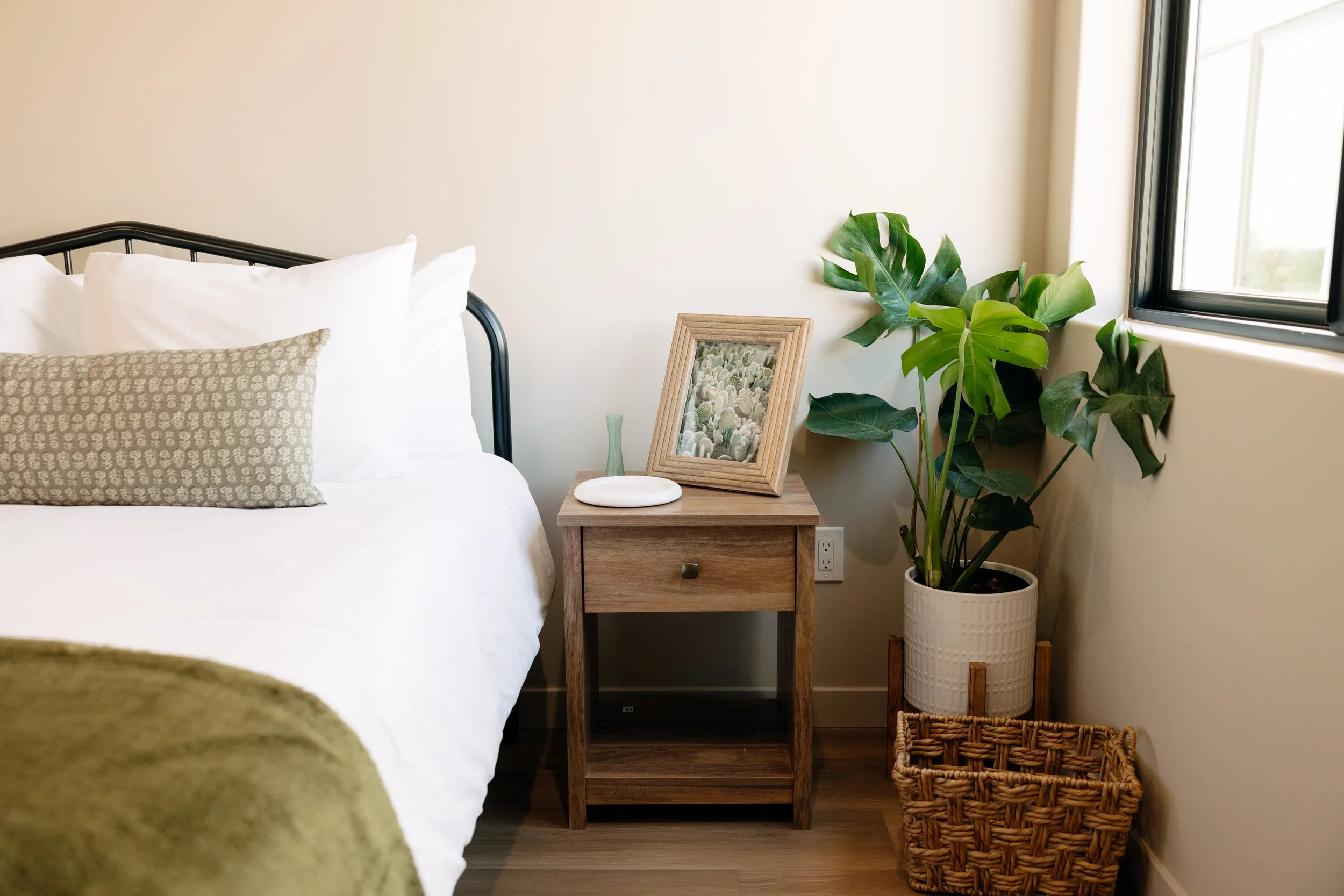 A bedroom corner with a bed, a wooden nightstand, a framed picture, a small green vase, a large potted plant, a woven basket, and a window.