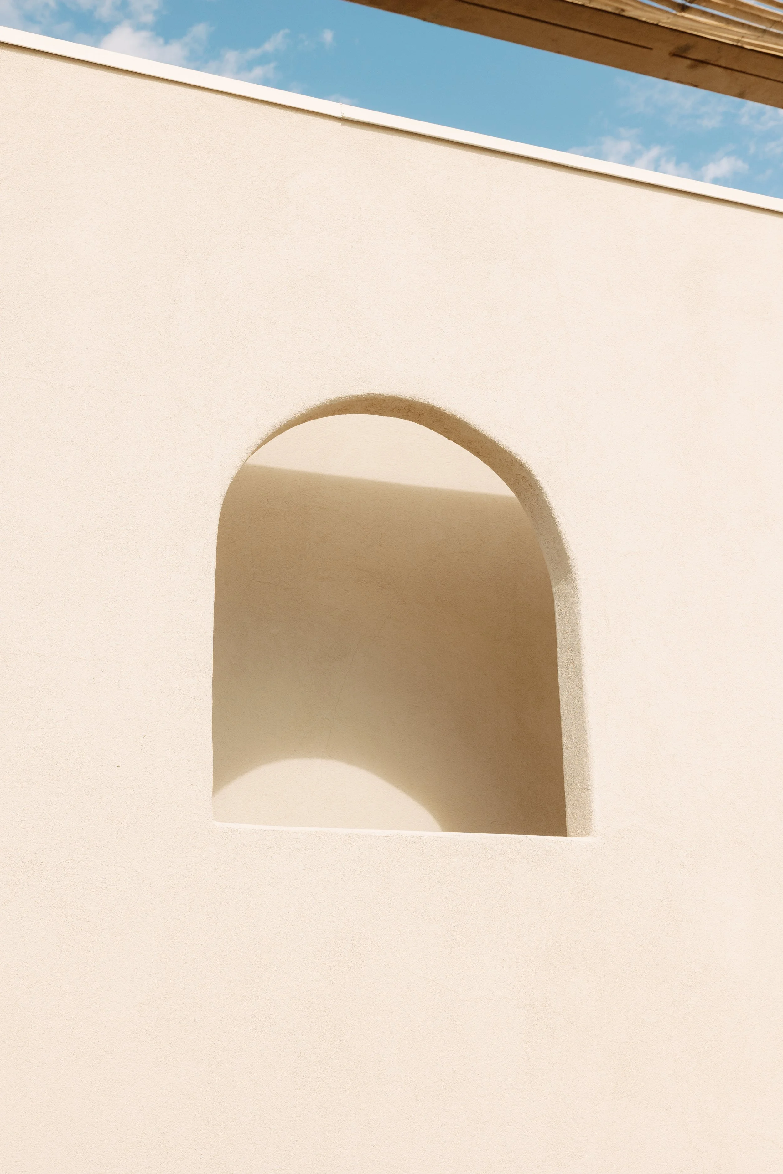 Close-up of a white wall with an arched opening and a shadow cast inside it, against a backdrop of a blue sky with scattered clouds.