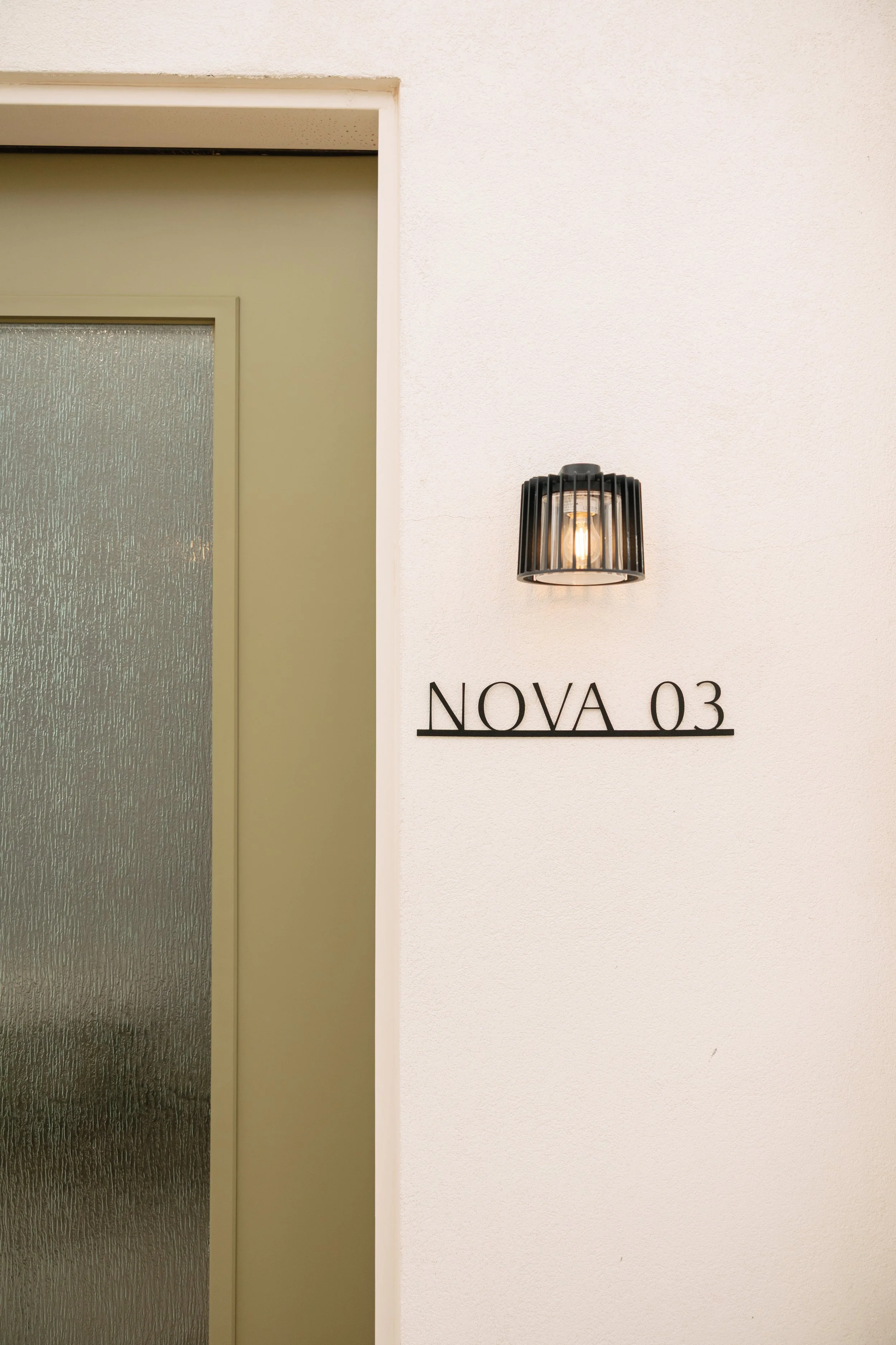 Interior wall with a black wall-mounted lamp, a frosted glass door, and the text 'NOVA 03' on the wall.