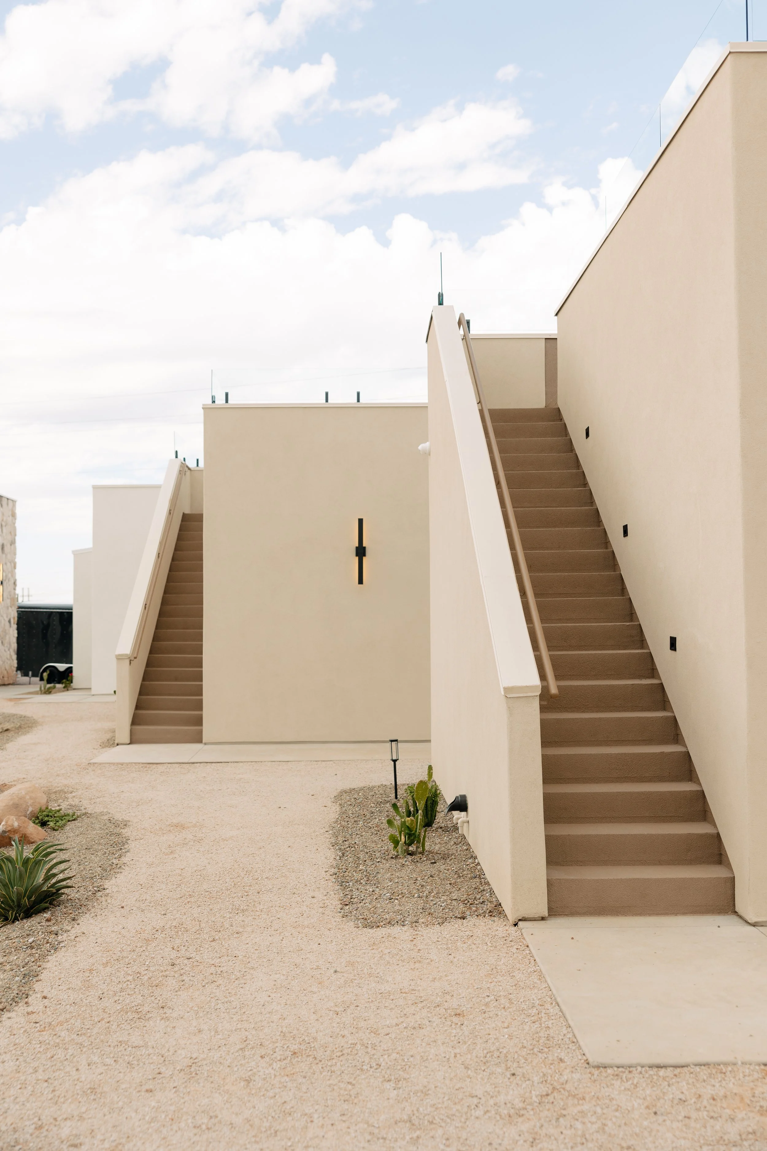 Two modern beige buildings with external staircases and handrails, set against a cloudy sky in a desert landscape.