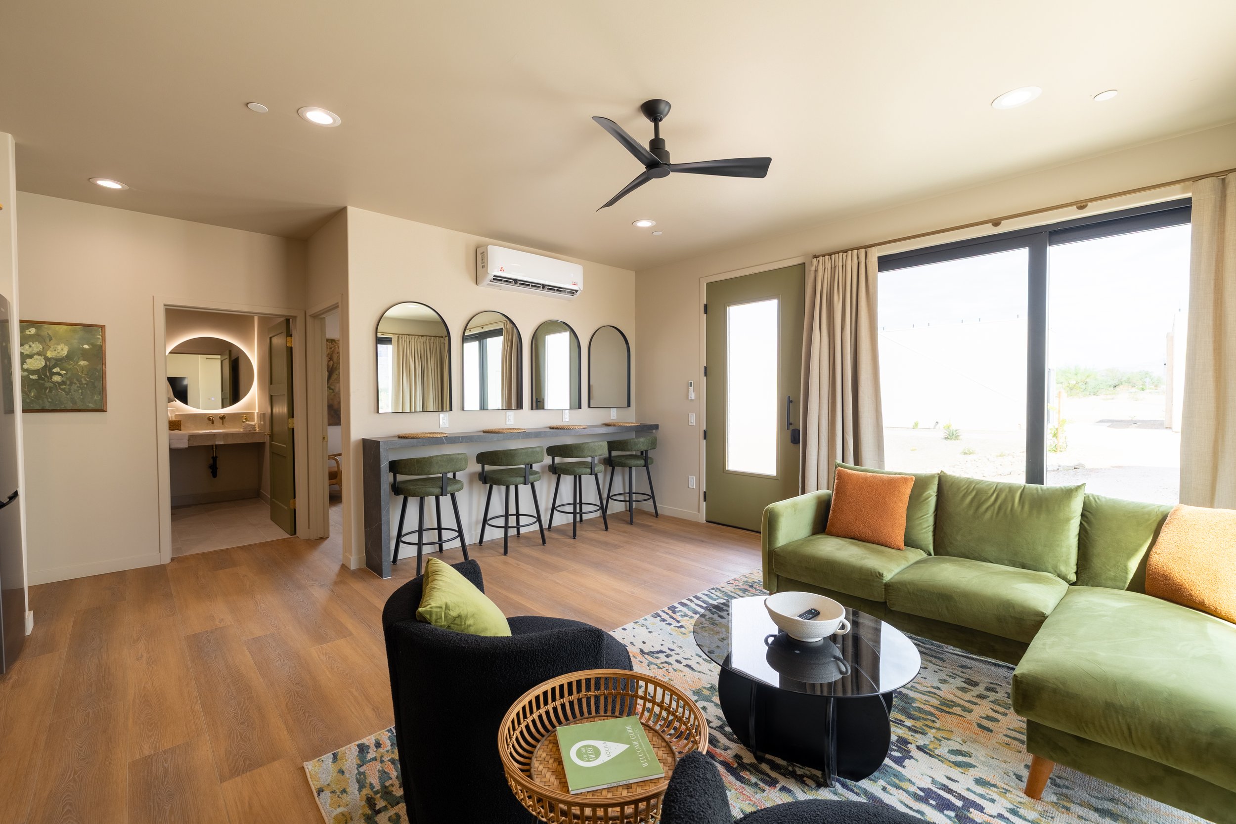 Living room with green sectional sofa, black and green armchair, glass coffee table, bar counter with stools, large window with curtains, doorway, and bathroom with mirror and lighting.