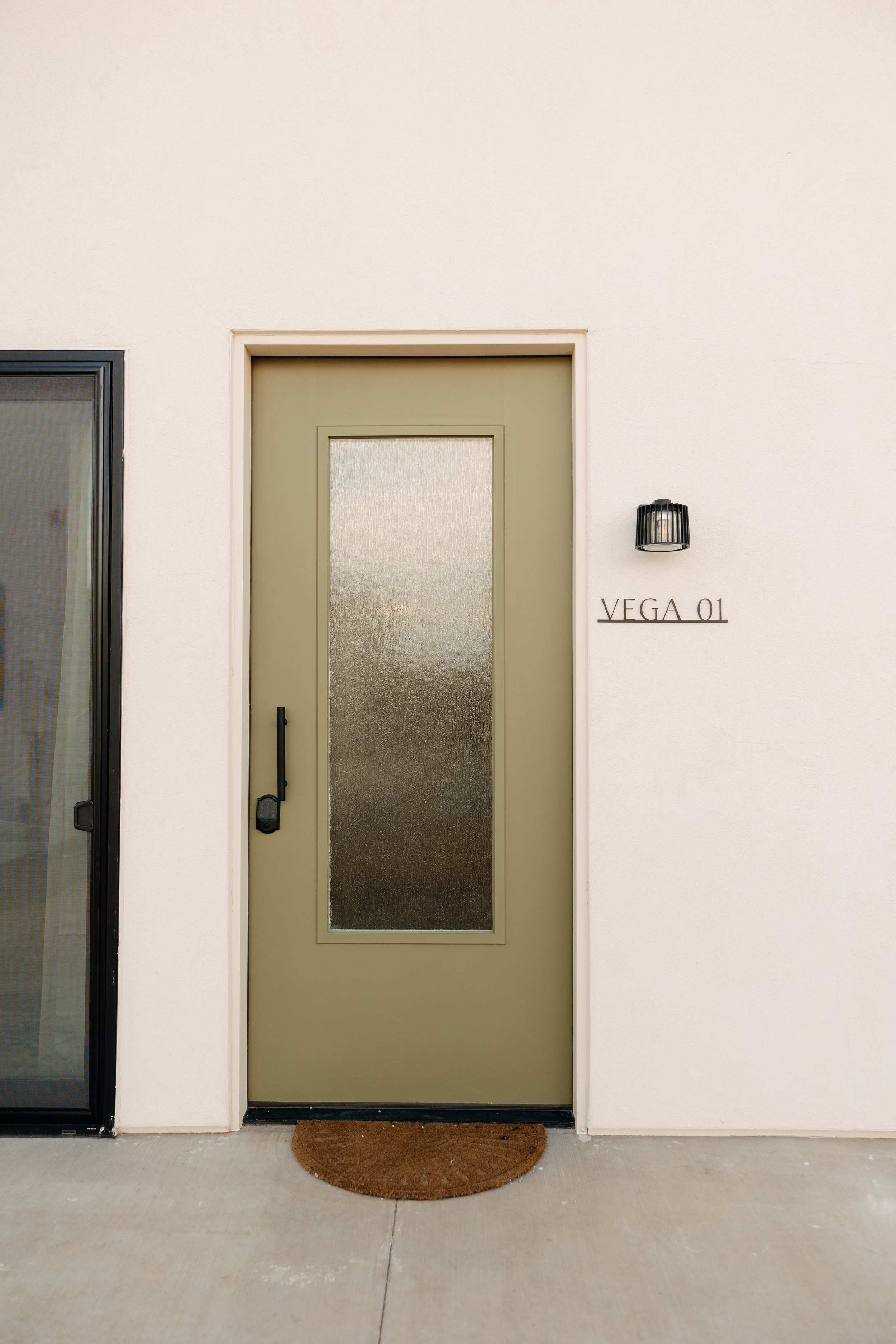 Apartment door with a textured glass panel, black handle, and a porch light on a white wall. Sign reads 'VEGA 01'.