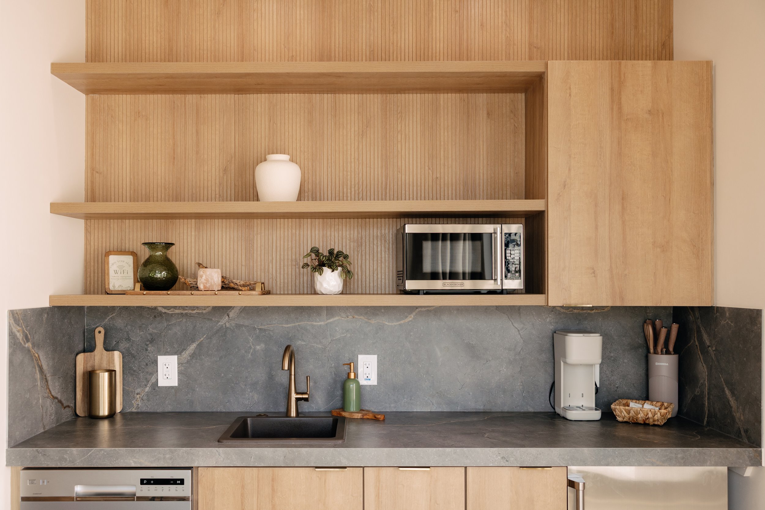 Modern kitchen with dark gray stone countertop, light wood cabinets, and open shelves. The counter has a gold and black container, a green soap dispenser, a coffee machine, and a basket of utensils. The open shelves contain a white vase, a dark green