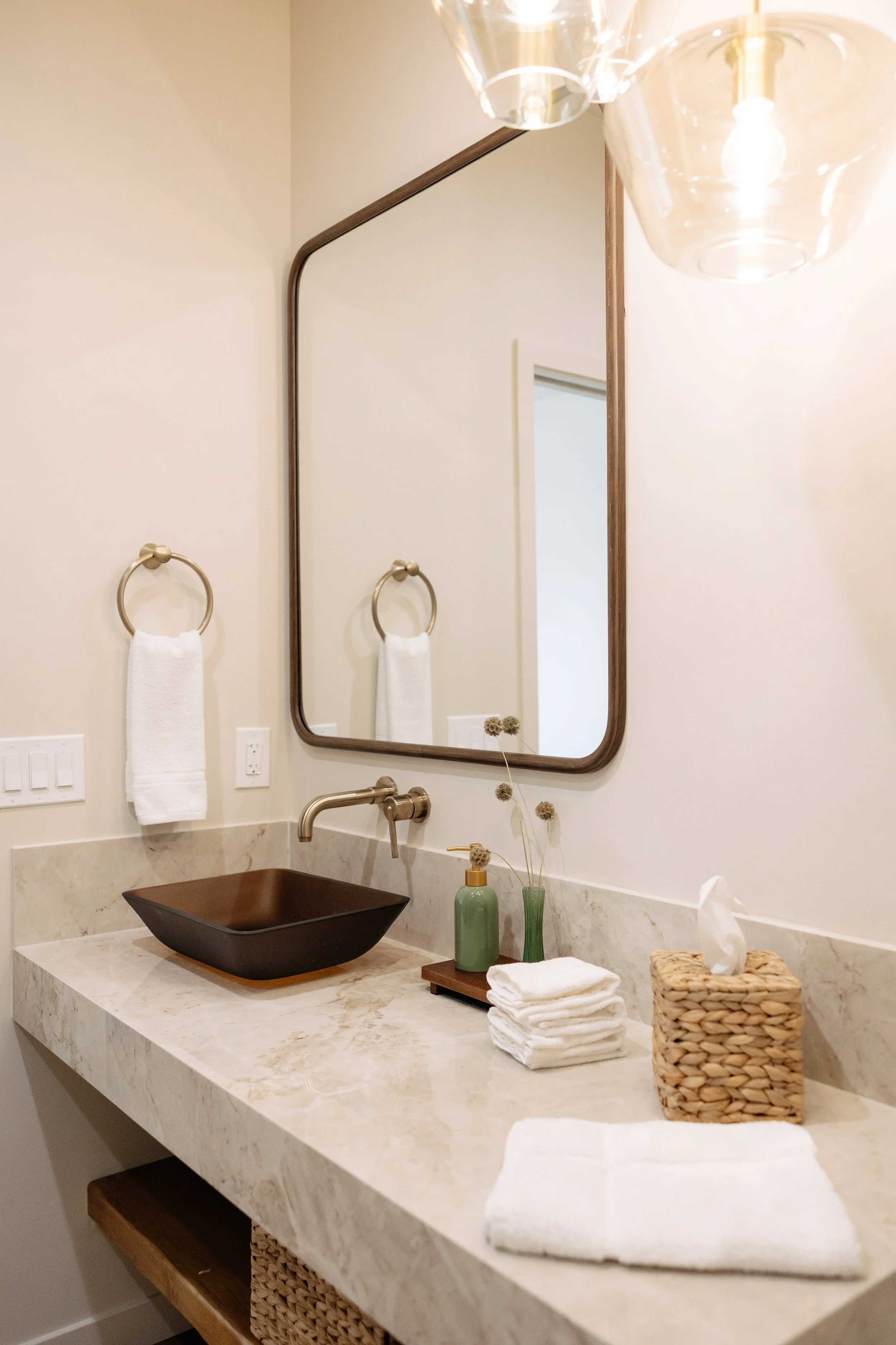 A bathroom vanity with a black vessel sink, a wall-mounted gold faucet, a mirror, a towel ring with a white towel, a green soap dispenser, a small vase with dried flowers, a stack of white towels, and a woven tissue box cover. There is a partial view