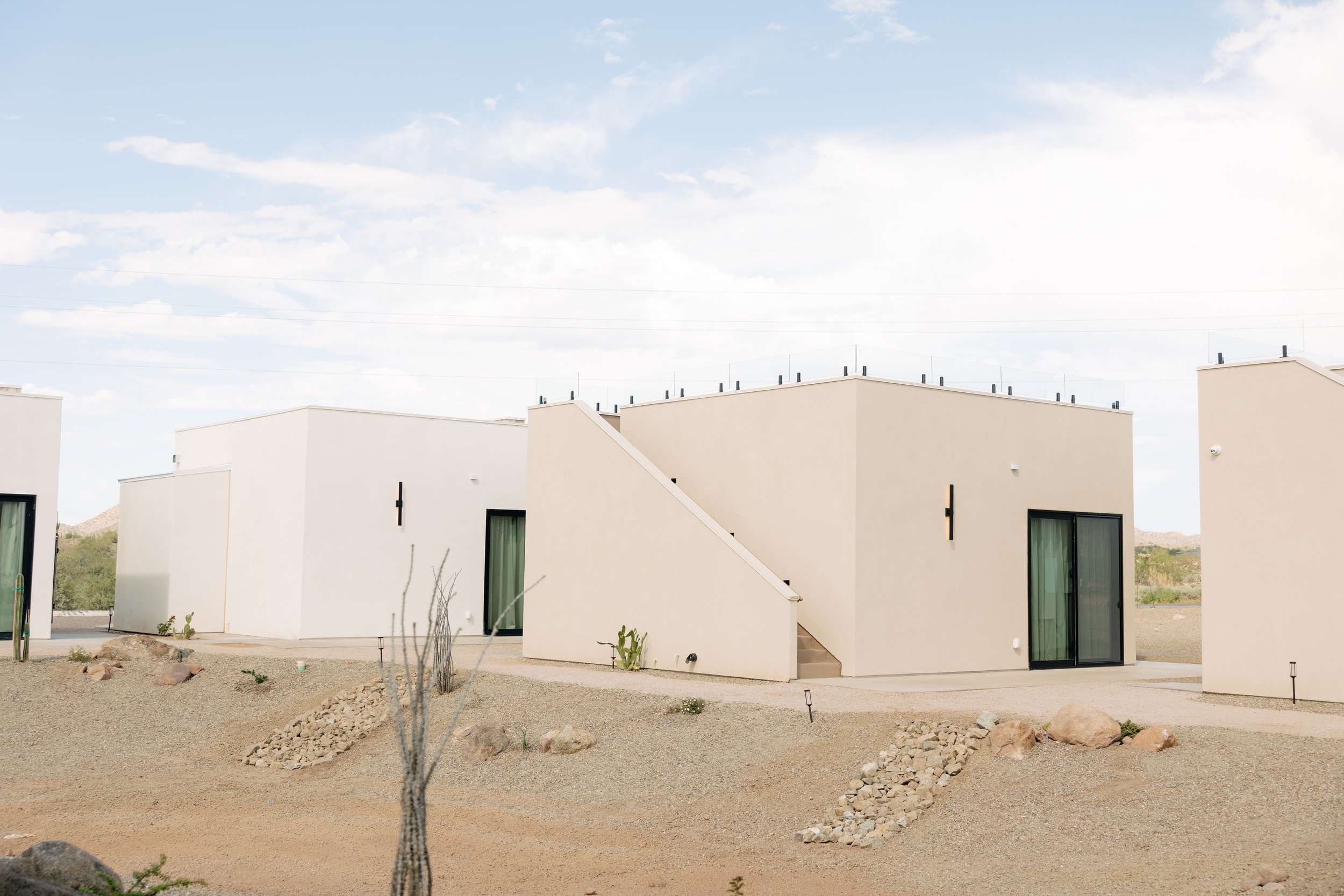 A row of modern, minimalist white houses in a desert landscape with a blue sky and scattered white clouds.