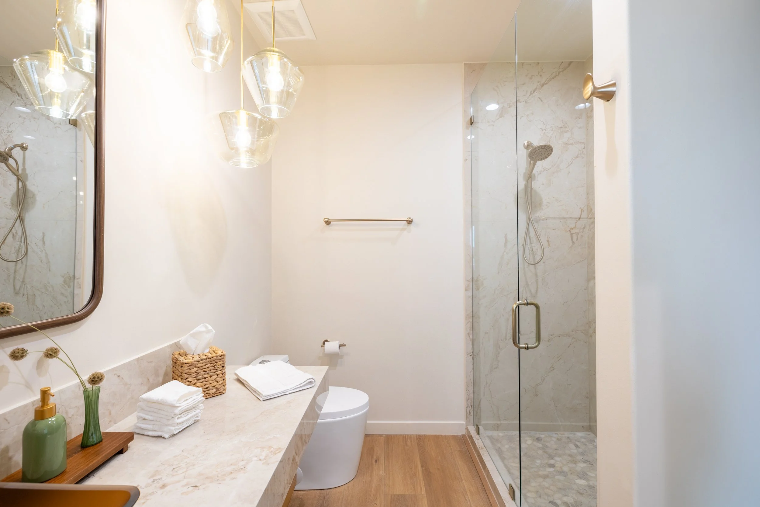 A modern bathroom with a glass shower, beige marble countertop, and minimalist decor. Features towels, a soap dispenser, and a mirror.