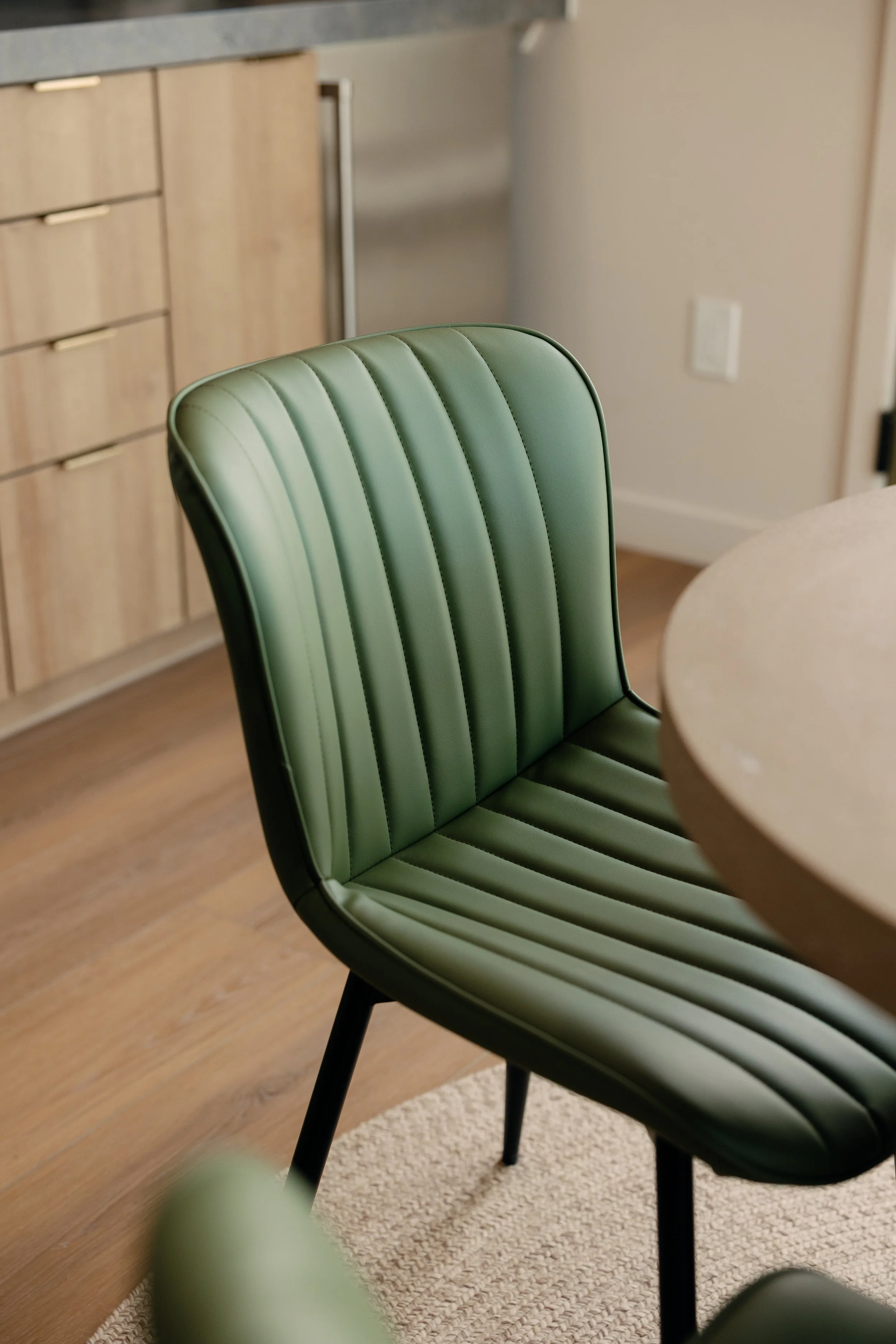 Close-up of a green upholstered dining chair with vertical stitching, positioned next to a round table in a modern kitchen or dining area.