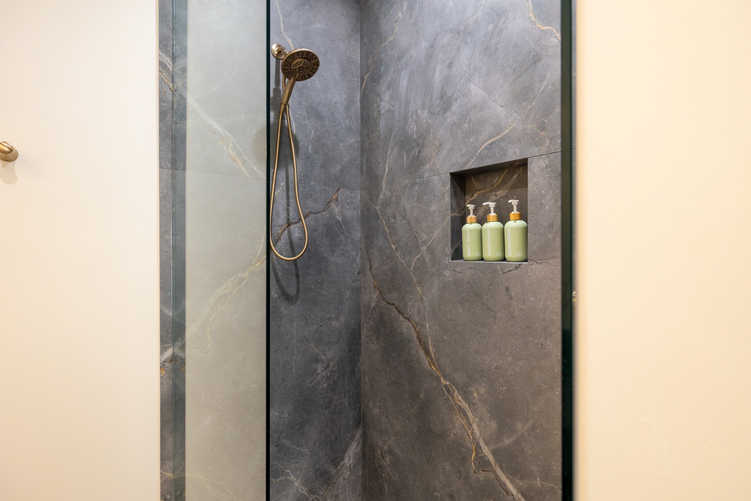 Shower with dark gray marble tiles, glass door, momentary showerhead, and built-in shelf with three green bottles labeled body wash, conditioner, and shampoo.