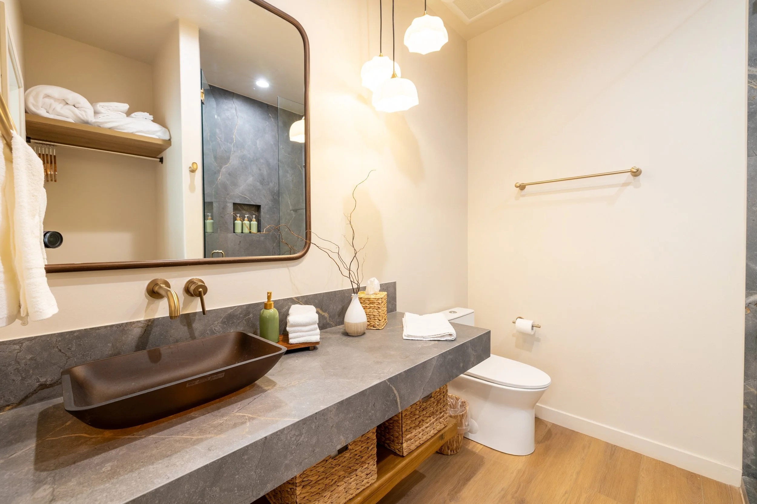 A modern bathroom with a large mirror, a dark vessel sink, and gold fixtures. There are rolled towels, a decorative branch, and a wicker basket on the counter. The room features a white toilet, a white wall, and warm lighting from hanging lamps.