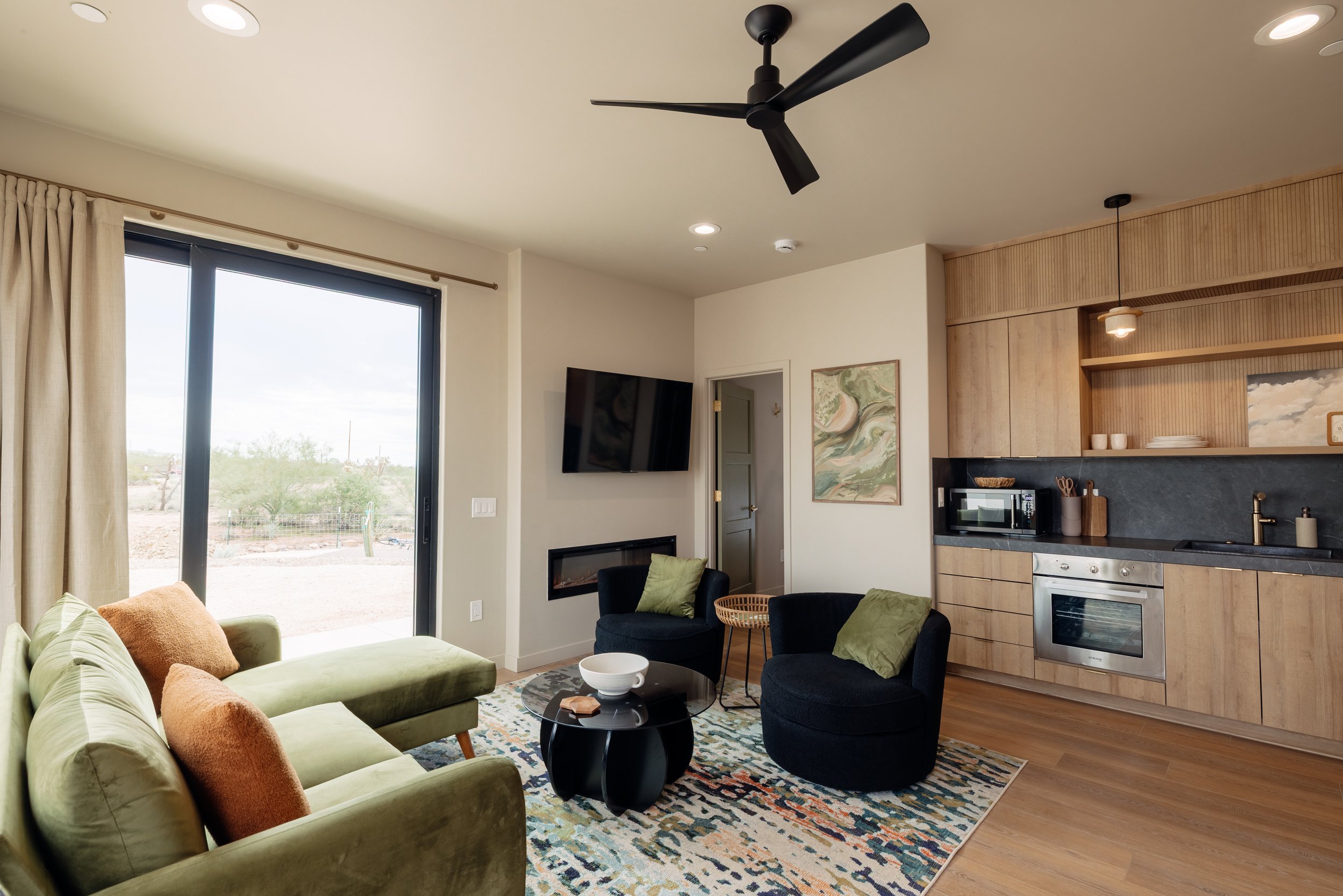 Living room with a green sofa, black armchairs with green pillows, coffee table, wall-mounted TV, fireplace, and kitchen area with wooden cabinets, black countertop, and built-in appliances. Large sliding glass door leads outside to a desert landscap