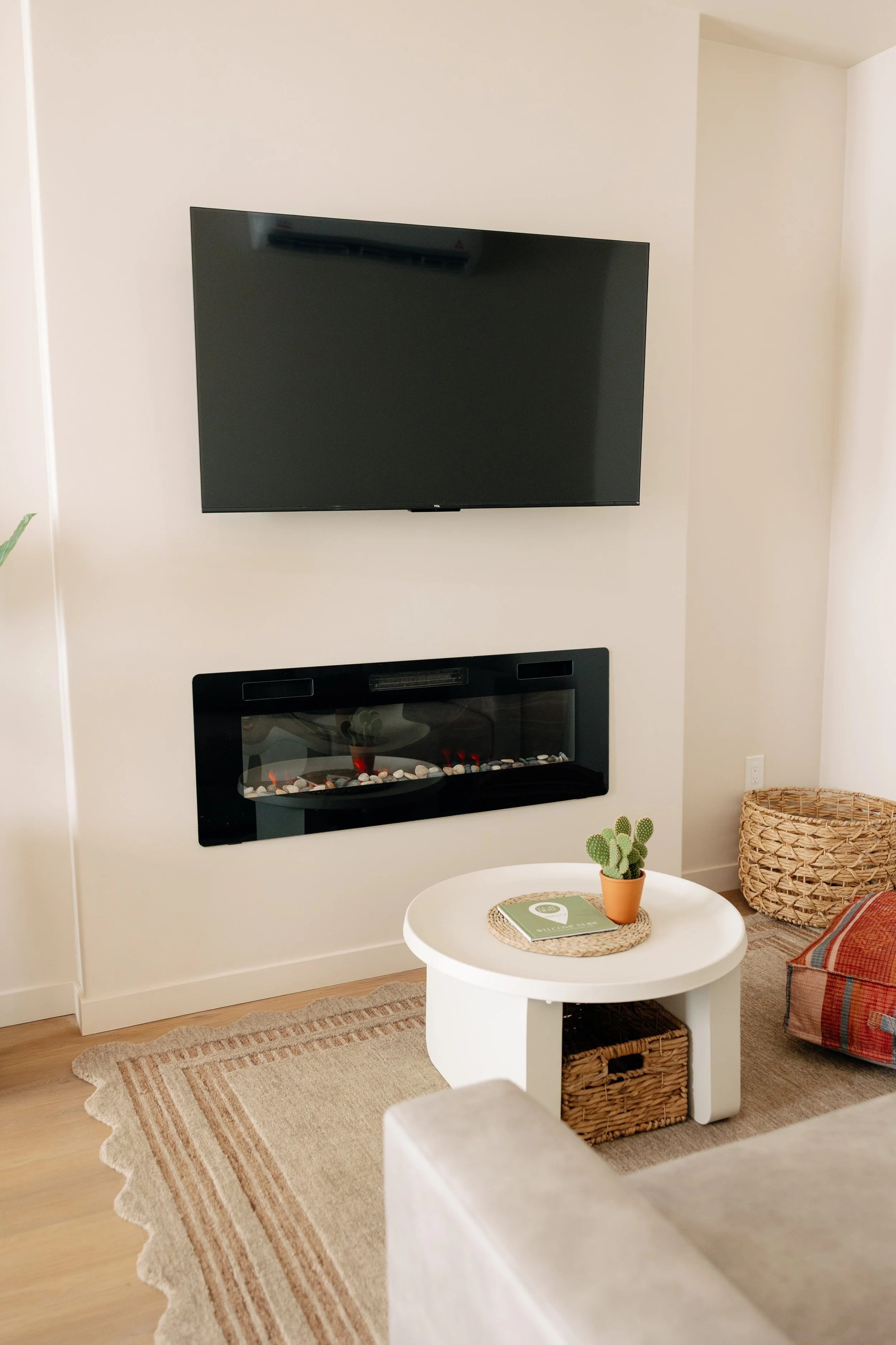 Living room corner with wall-mounted TV, electric fireplace, white round coffee table with a small potted cactus, woven basket, and a beige and striped rug.