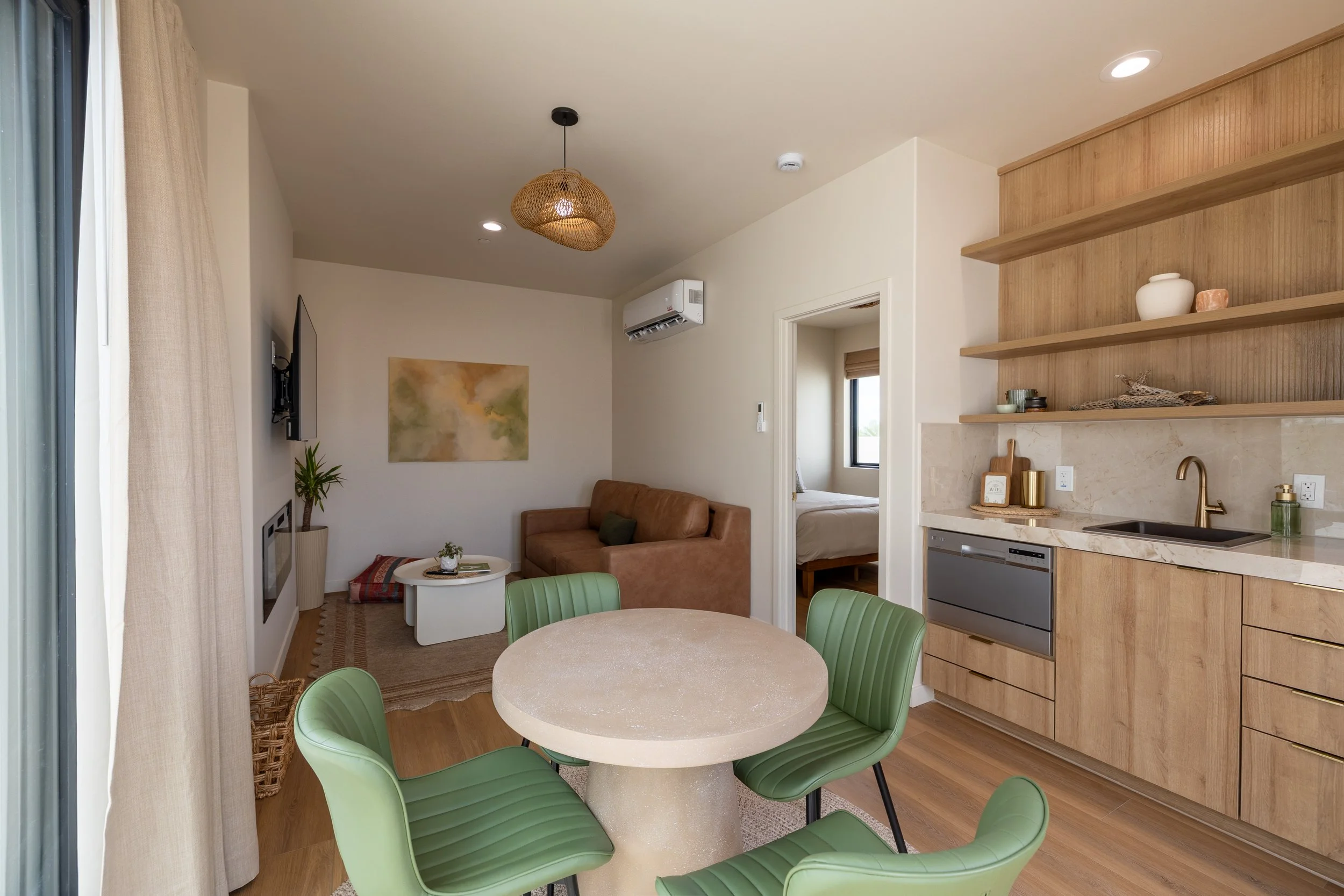 Interior of a modern apartment with a small kitchen, a dining table with green chairs, and a living area with a brown sofa, a wall-mounted TV, and a window with beige curtains.