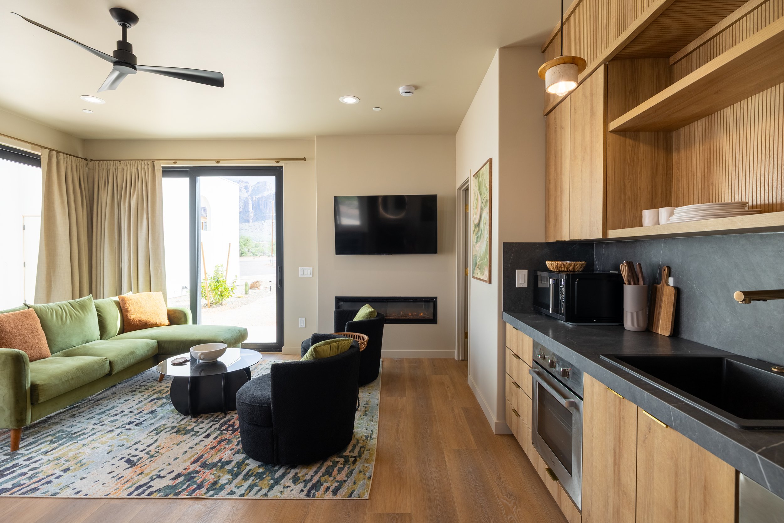 Living room with green sofa, black chairs, sliding door, mounted TV, fireplace, kitchen area with microwave, plates, and black countertop.