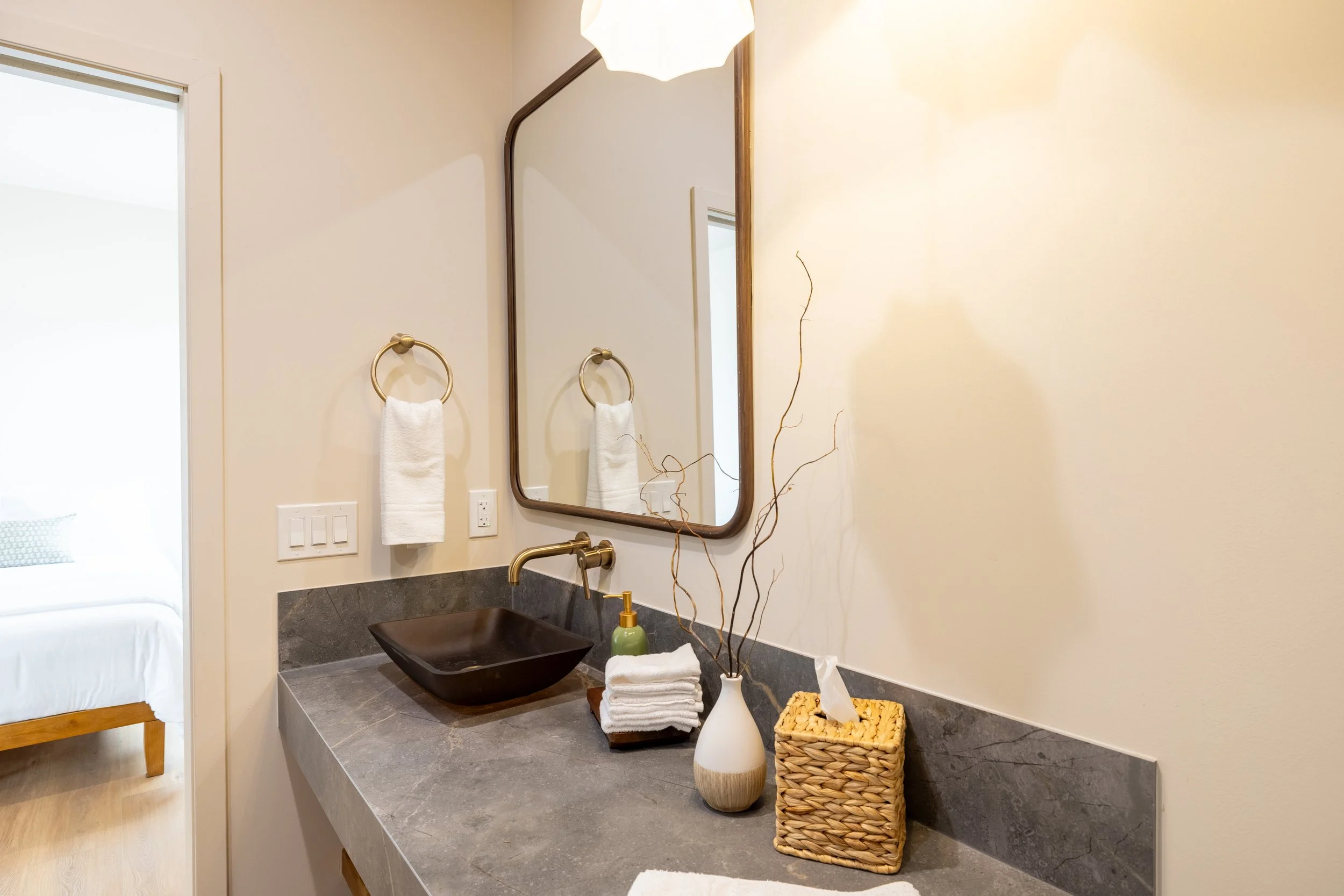 Bathroom sink with a black vessel basin, gold faucet, mirror, towel ring with a white hand towel, wall switch, decorative vase with dry branches, soap dispenser, tissue box, and an arm of a bedroom visible through the doorway.