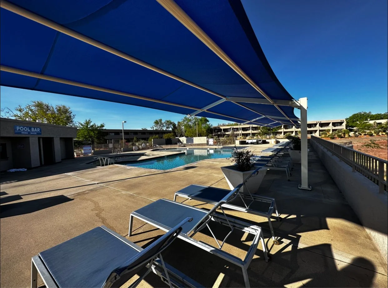 Empty swimming pool area with lounge chairs, shaded by a large blue awning, near a building with a sign that reads 'Pool Bar' on a sunny day. Commercial shade sails. commercial shade awning, shade canopy, shade canopies, shade umbrella.